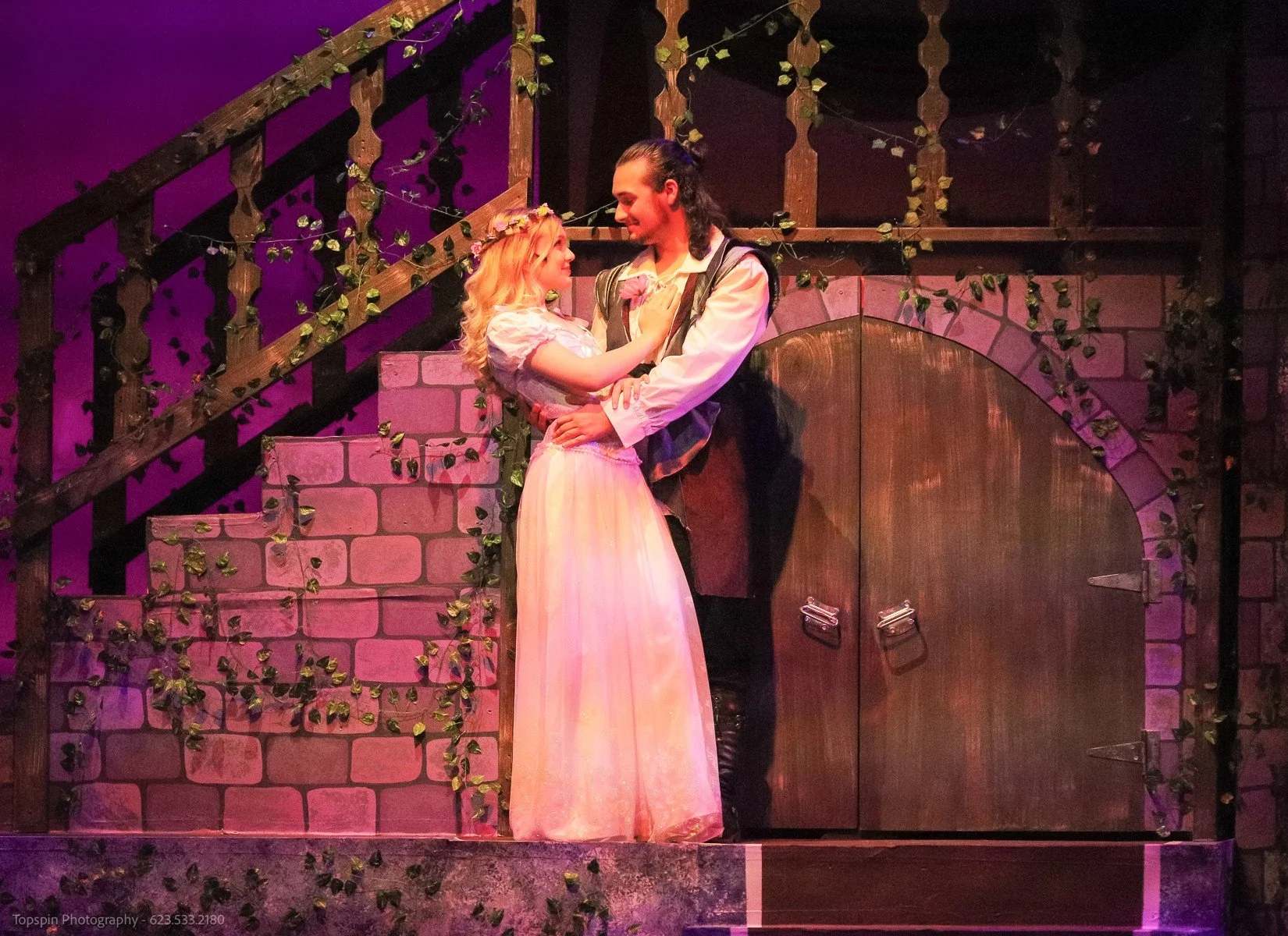 A male and female actor in period costumes sharing an intimate moment on stage, with a set designed to resemble a castle or medieval structure, decorated with vines and bricks.