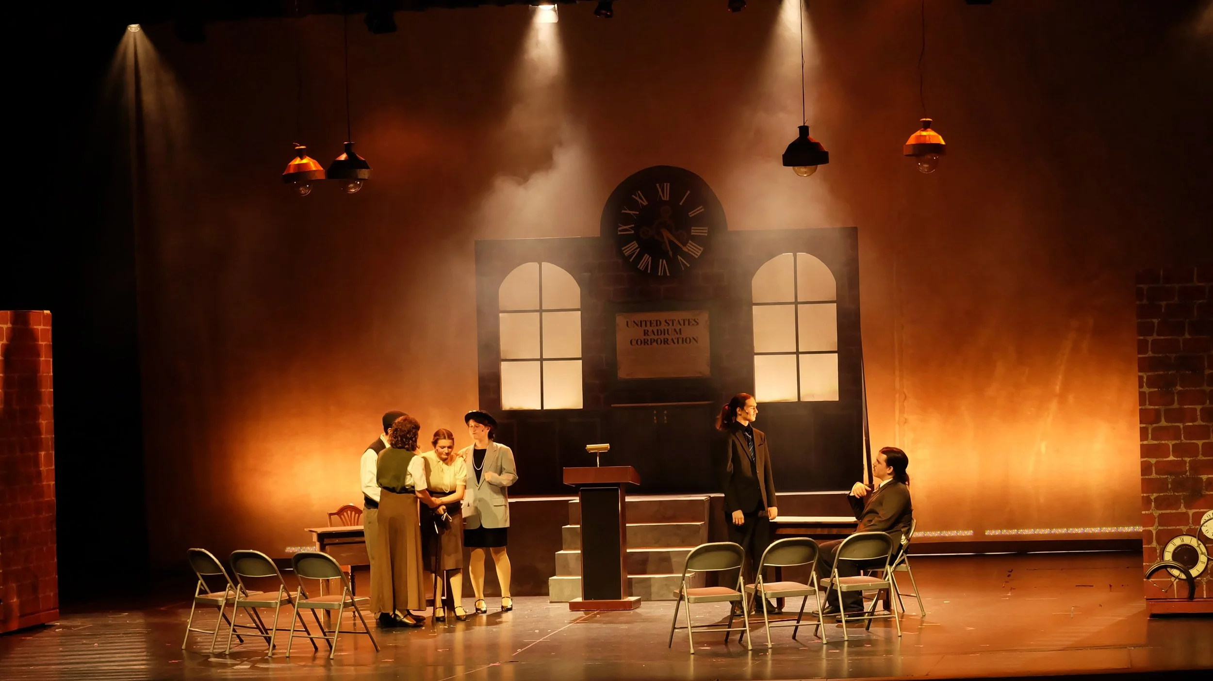 A theatrical stage set resembling an office with a clock and large windows backdrop, with actors in period costumes engaged in conversation, and chairs arranged in the foreground.