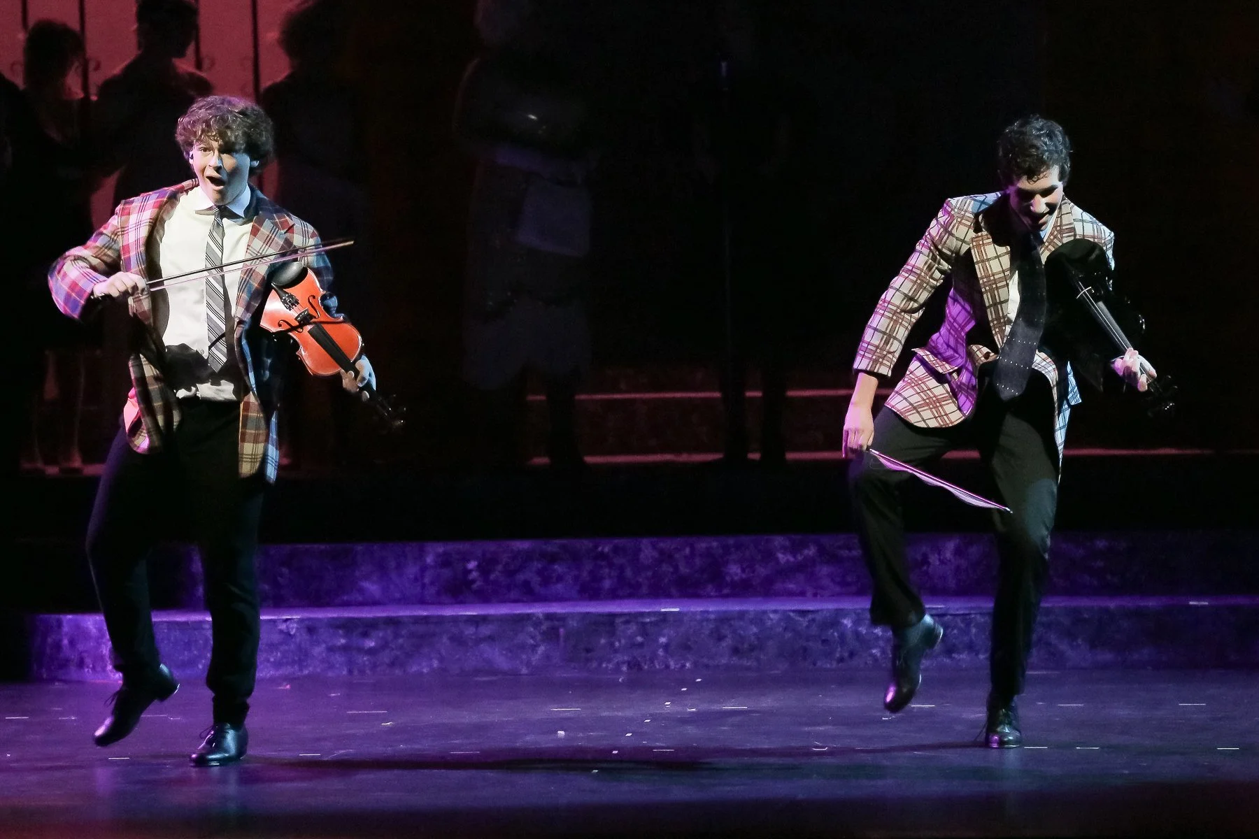 Two male performers dressed in plaid blazers and dark pants are on stage, playing violins and dancing. They are illuminated with stage lighting, with dark silhouettes of other performers in the background.
