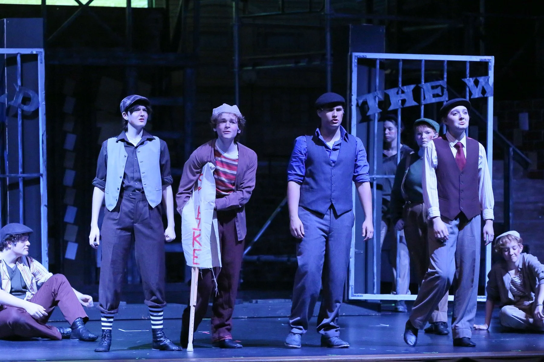 The image shows a theatrical stage with six young actors dressed in vintage clothing, standing and sitting in various poses. The background features a metal fence with a sign that reads 'THEY.' The scene appears to be part of a play set during a hist