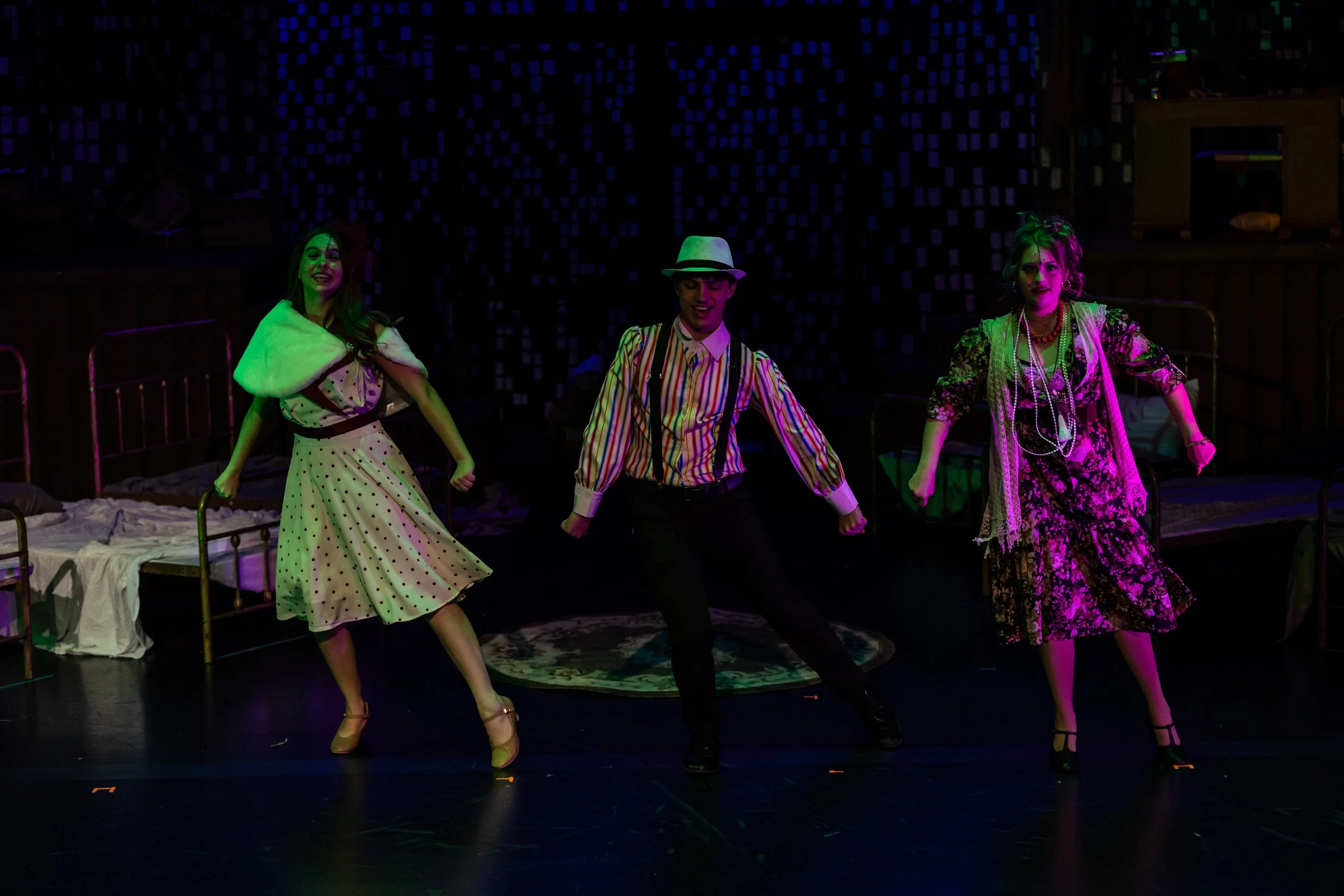 Three performers dressed in vintage clothing dancing on a stage with a dark background and colorful lighting.