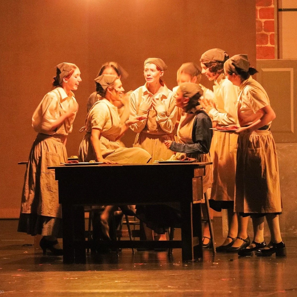 Group of women dressed in historical uniforms, gathered around a desk on stage, likely performing a play or musical.