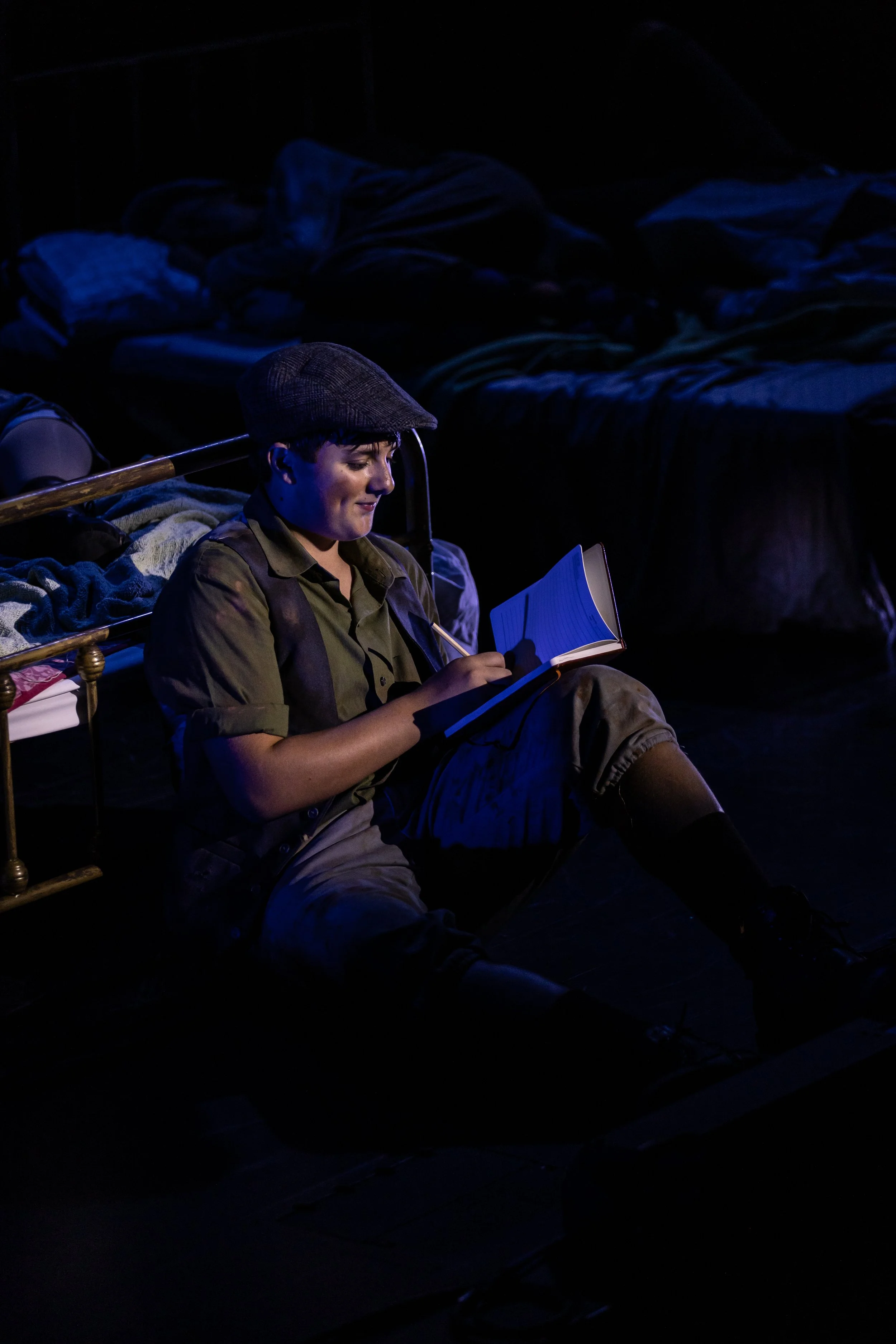 Young man sitting on the floor, reading a book in a dark room with a bed in the background.