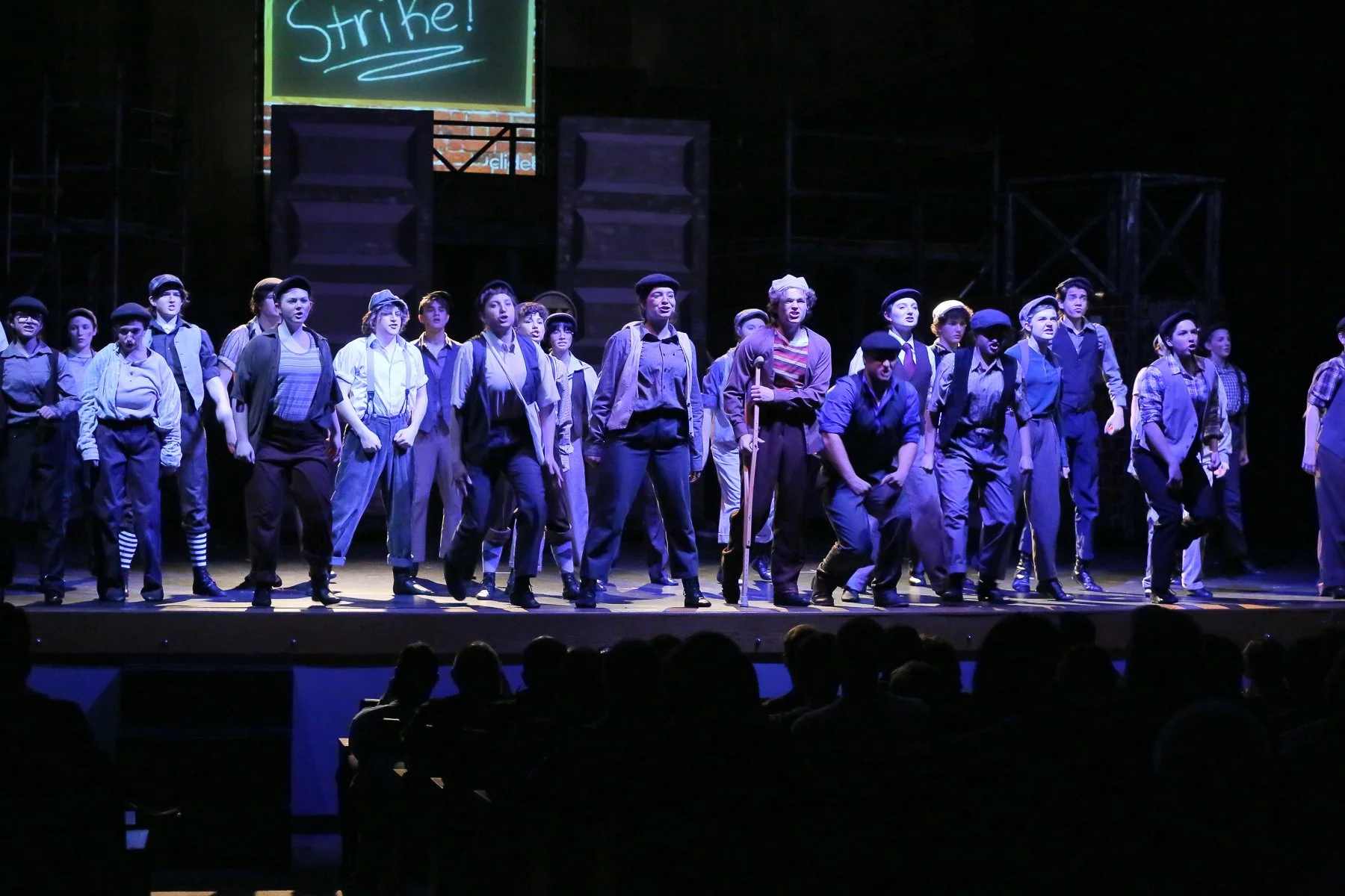 A group of actors dressed in historical worker costumes perform on a stage in front of an audience. The scene is dark with a spotlight highlighting the performers, and there's a large screen above displaying the word 'Strike!' in bright letters.