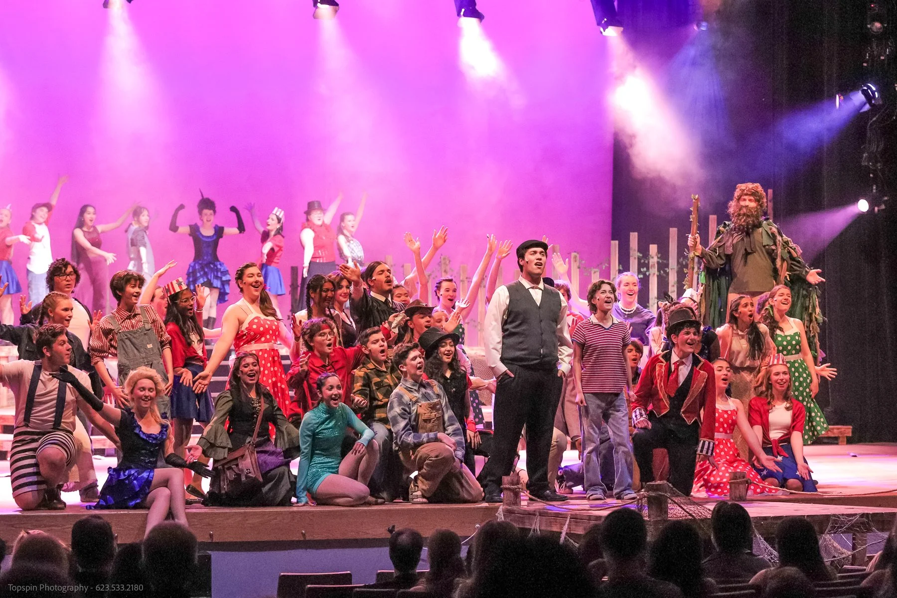 A large group of performers on stage, dressed in colorful costumes, singing and dancing with a pink and purple backdrop, with stage lights shining down. Some characters are dressed as children, while others represent animals and fantastical figures.
