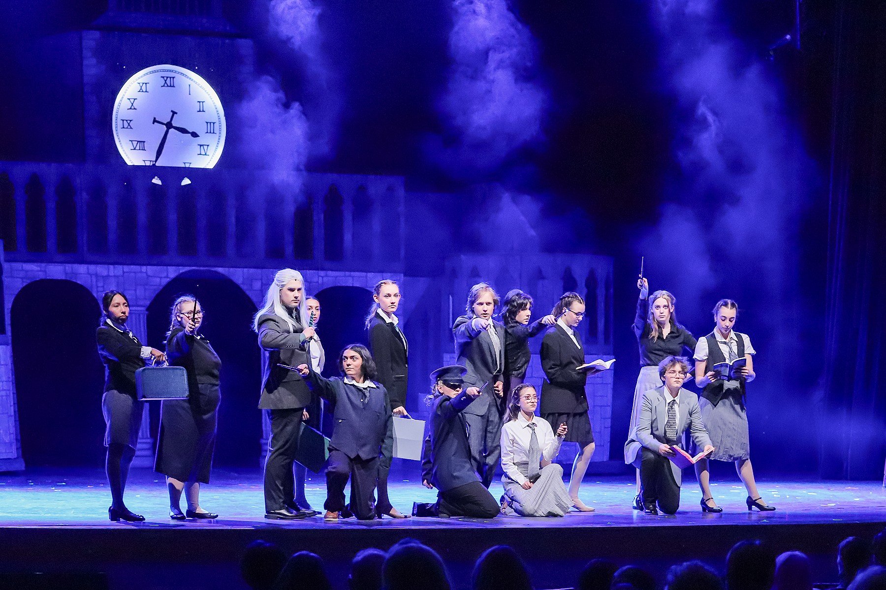 The image shows a theatrical stage performance with a group of actors dressed in formal and costume clothing, some holding books or props, in front of a backdrop depicting a large clock and a building with arches, under blue lighting and smoke effect