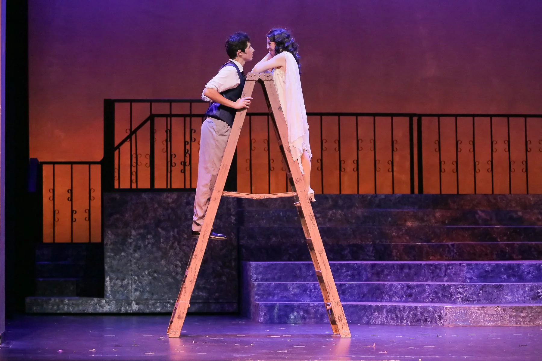 A scene from a theatrical performance showing a man and a woman standing on a ladder on stage. The man is leaning on the ladder holding onto it with one hand and facing the woman, who is touching her face with one hand. The stage has a background wit