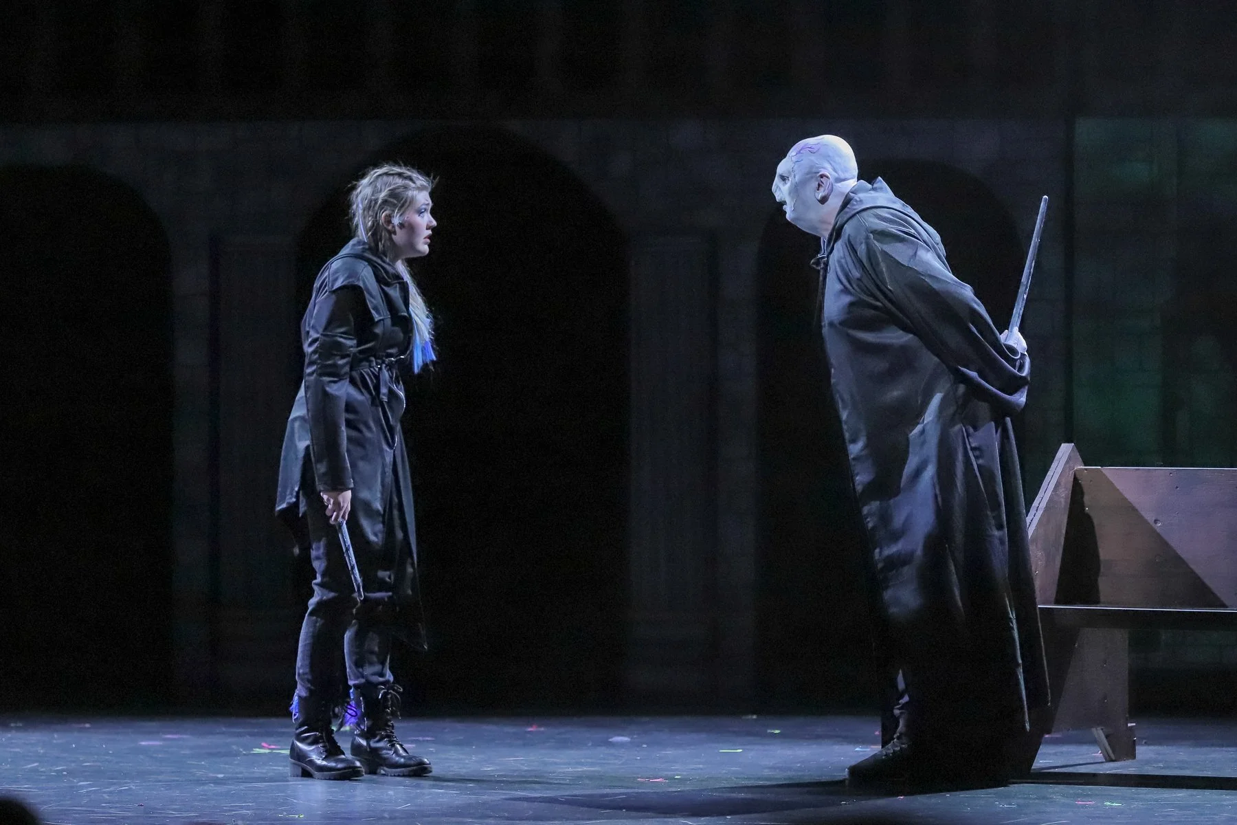Two actors in black costumes face each other on stage. The woman on the left holds a gun, and the man on the right, dressed as a clown with a broom behind him, leans forward. The scene is dark with a theatrical setting.