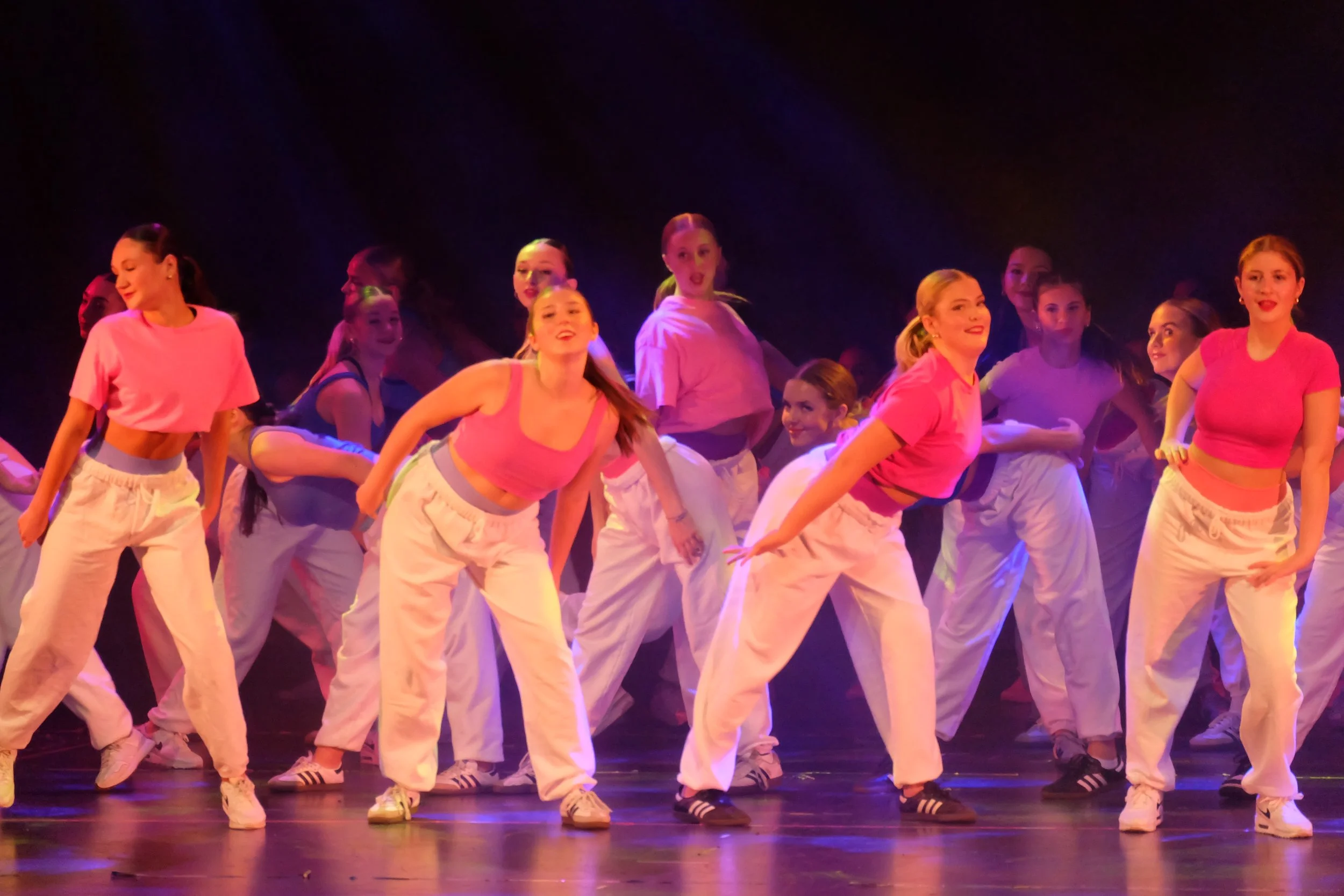 Group of dancers on stage wearing pink tops and white pants performing a dance routine.