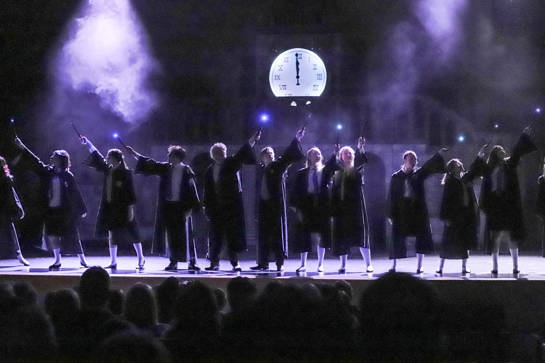 A group of people dressed in dark robes standing on stage, holding wands with glowing tips, performing a magic show with a large clock in the background showing nearly midnight.