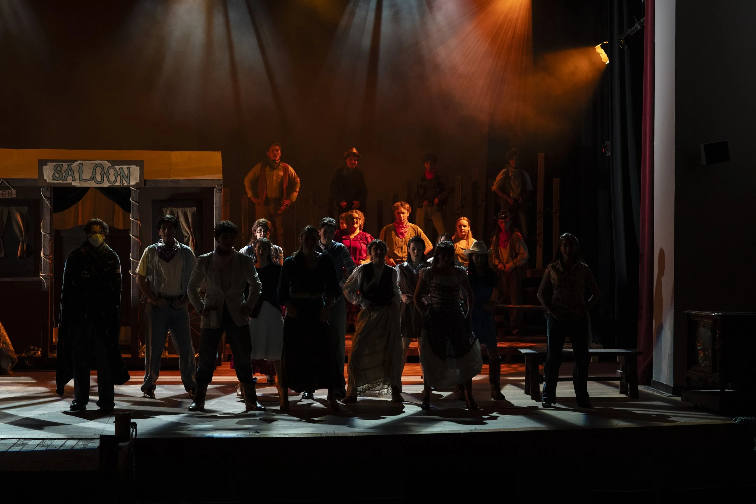 Stage scene with actors dressed in old-fashioned Western costumes, some standing and some walking, with a saloon set, dim lighting, and backdrop suggesting a Western town.