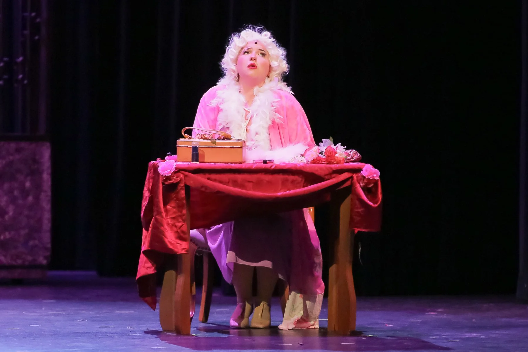A woman with curly blonde hair is sitting at a table on a stage, wearing a pink robe with a white feather boa. She has an expressive look, possibly acting or singing, with a small box, lipstick, and flowers on the table. The background is dark with s