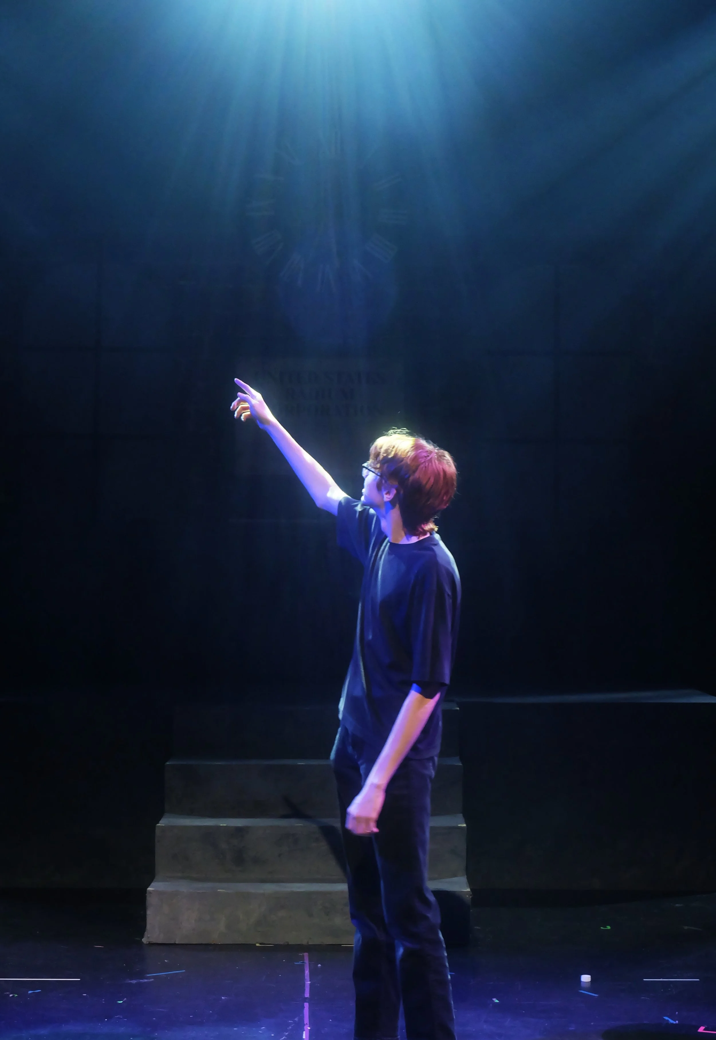 A person with reddish-brown hair, wearing glasses and dark clothing, standing on a dark stage with steps, reaching upward with one arm, illuminated by blue stage lighting.