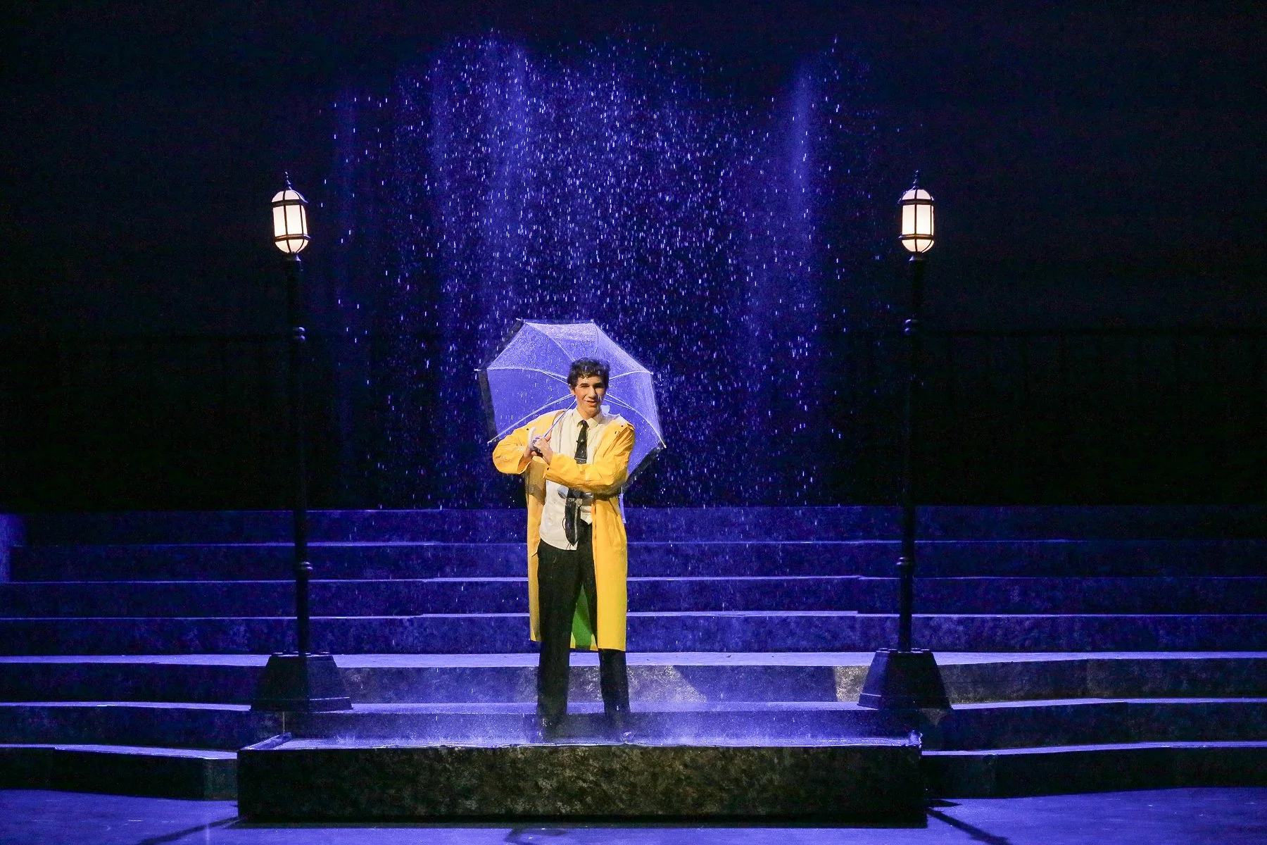 A man standing on a stage in the rain, holding a transparent umbrella, flanked by two street lamps, with steps behind him, during a theatrical performance.