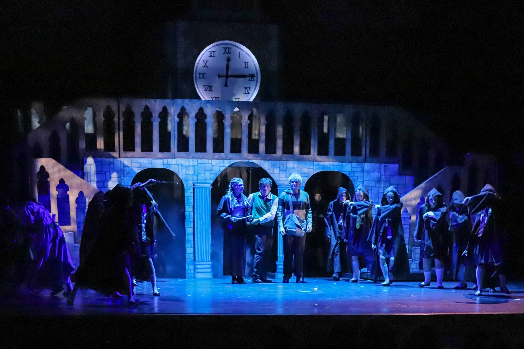 Stage scene from a theatrical play with performers dressed in costumes, set against a backdrop resembling a castle with a clock showing 3:15.