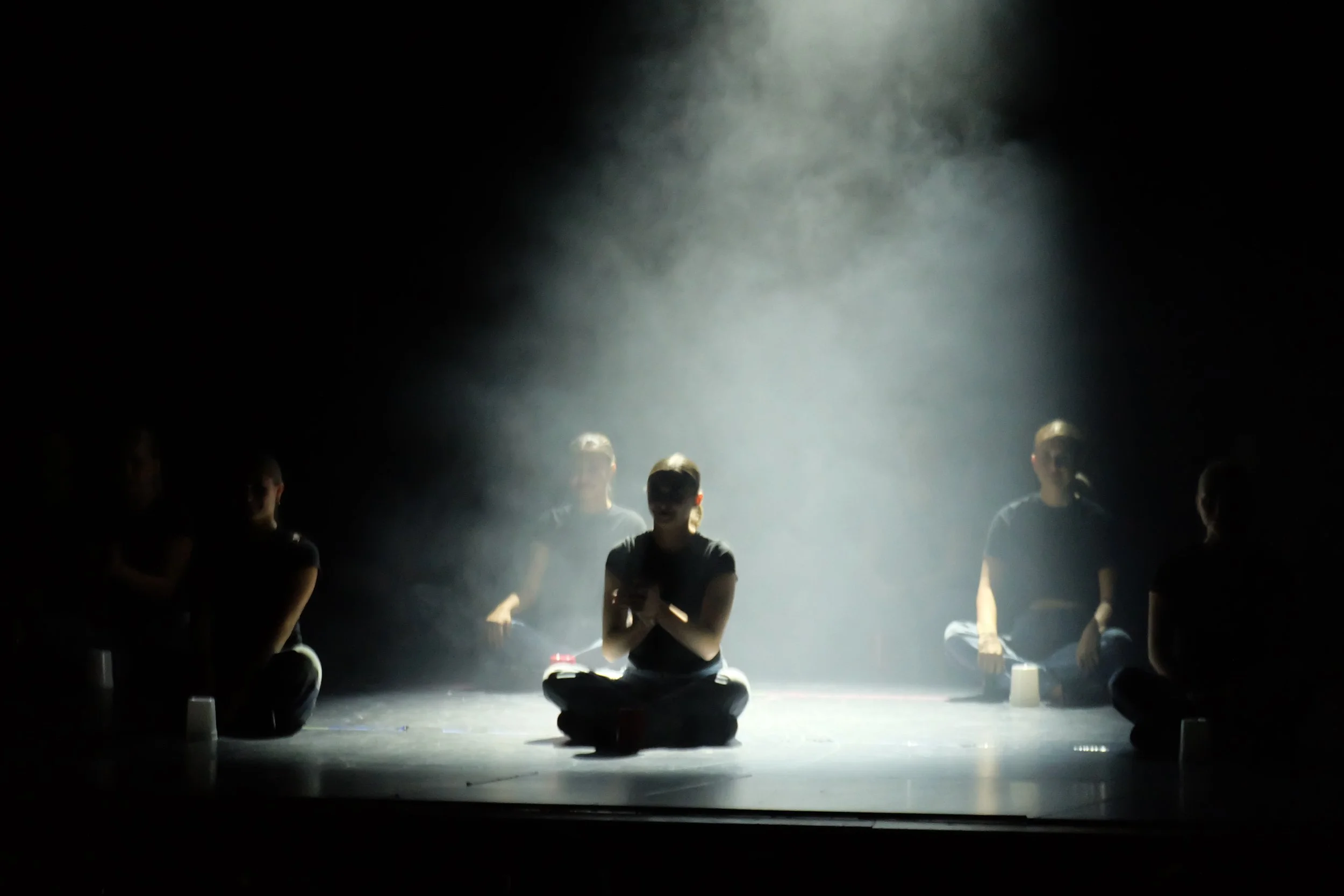 Dimly lit stage with several people sitting cross-legged, some with their eyes closed, surrounded by fog and a spotlight shining down.