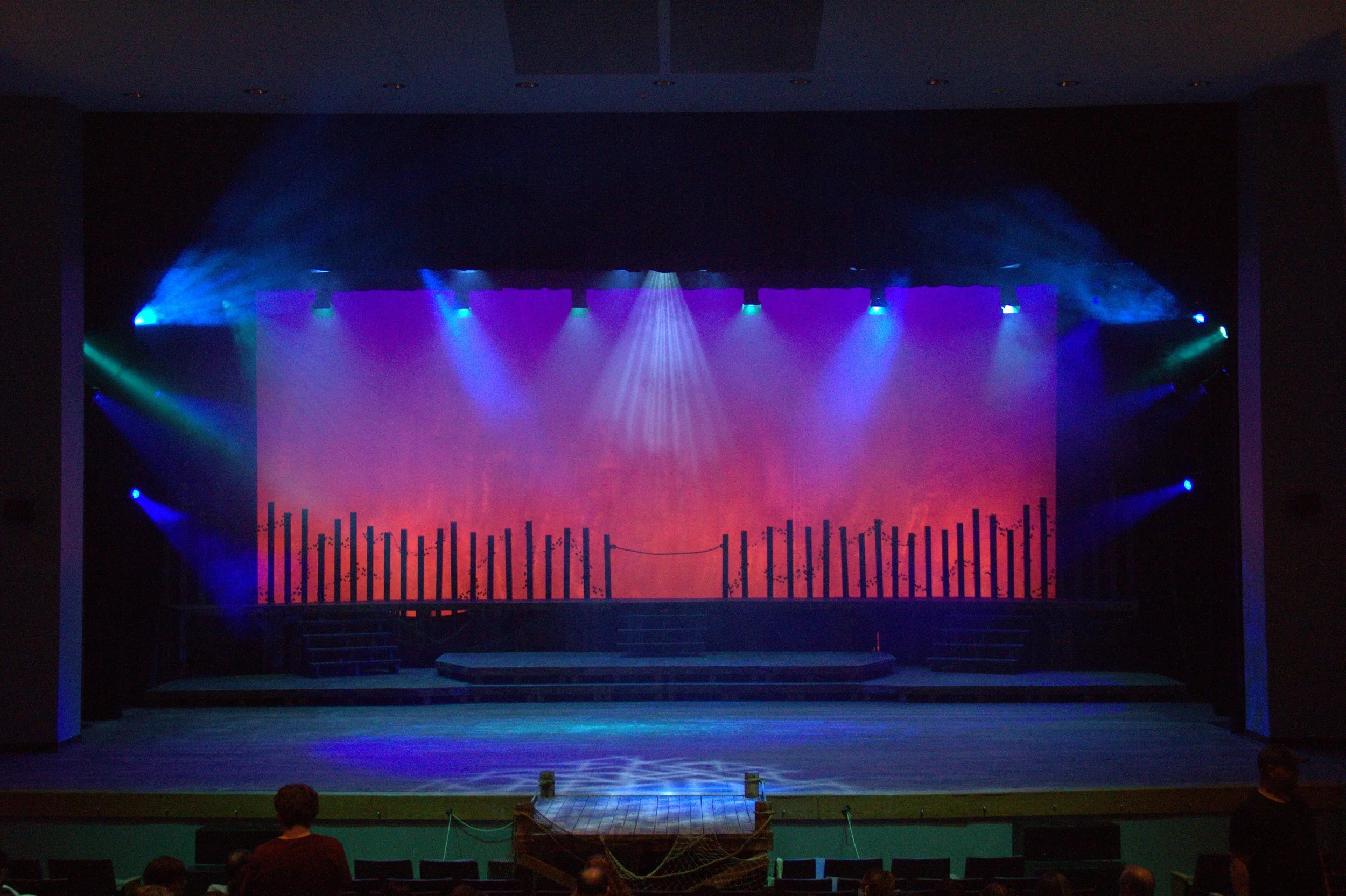 Empty theater stage with colorful spotlights and light fog, audience seating visible at the bottom.
