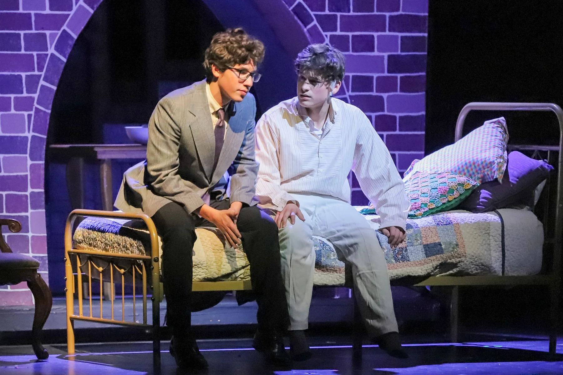 Two young men sit on a bed in a dimly lit room with purple lighting. They appear to be engaged in a serious conversation, with one in a suit and the other in casual pajamas. The room has a brick wall backdrop and a small table next to the bed.