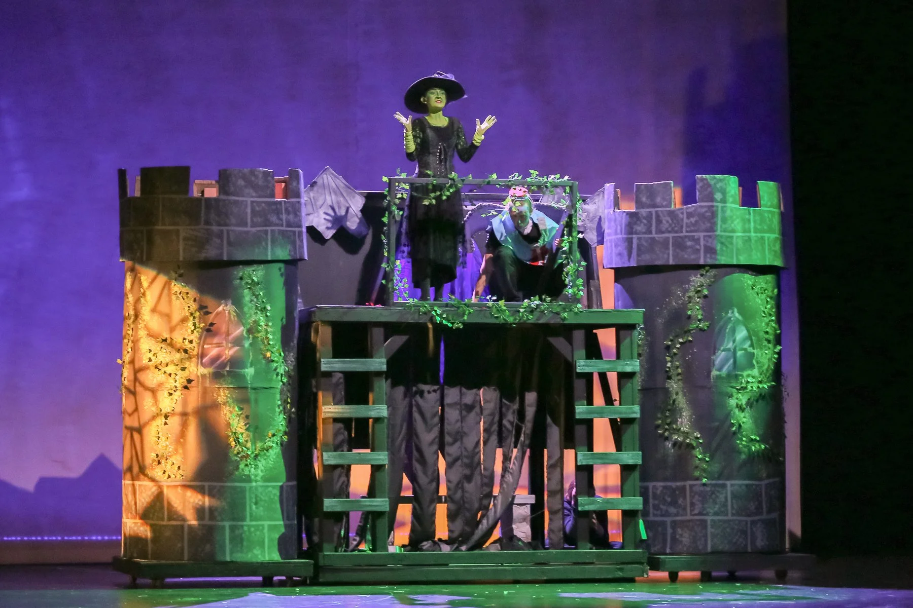 A theatrical stage set resembling a castle with two women in costumes, one in black with a wide-brimmed hat and green face paint, and the other with a headband of flowers. The scene features castle towers with greenery and a purple background.
