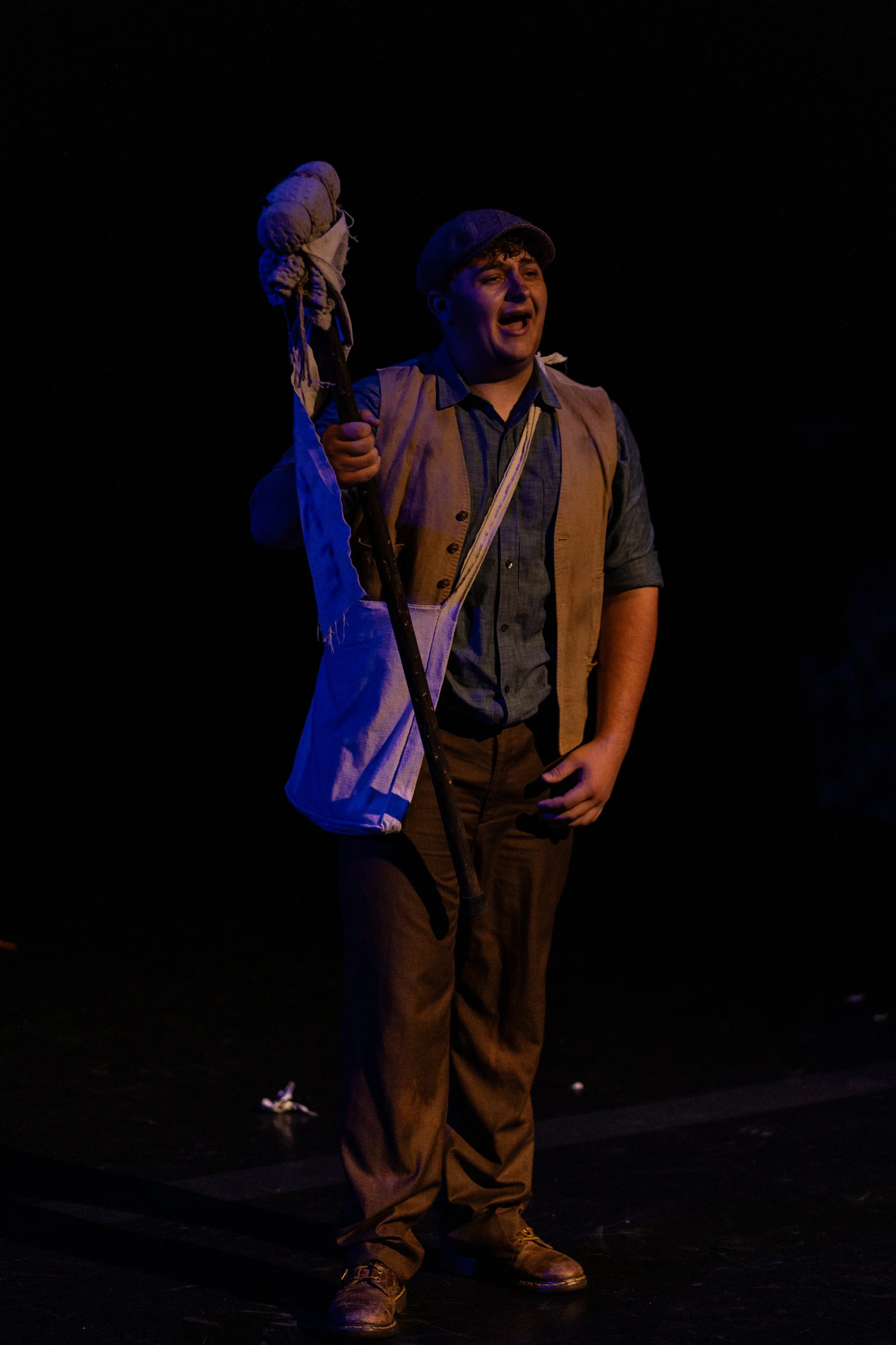 A young man performing on stage, dressed in vintage clothing with a newsboy cap, holding a makeshift staff or cane, with a dark background.