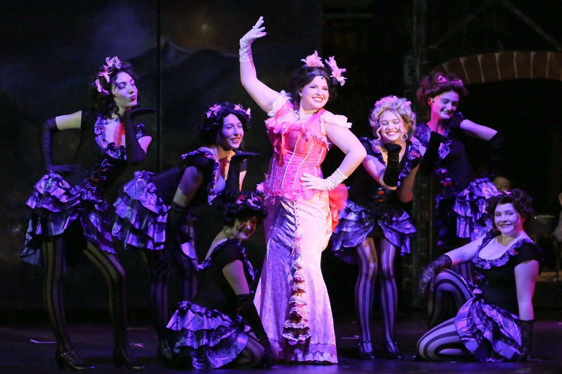 Stage performance with women in vintage costumes, one in a pink dress and the others in black and purple dresses, posing with smiles and hand gestures.