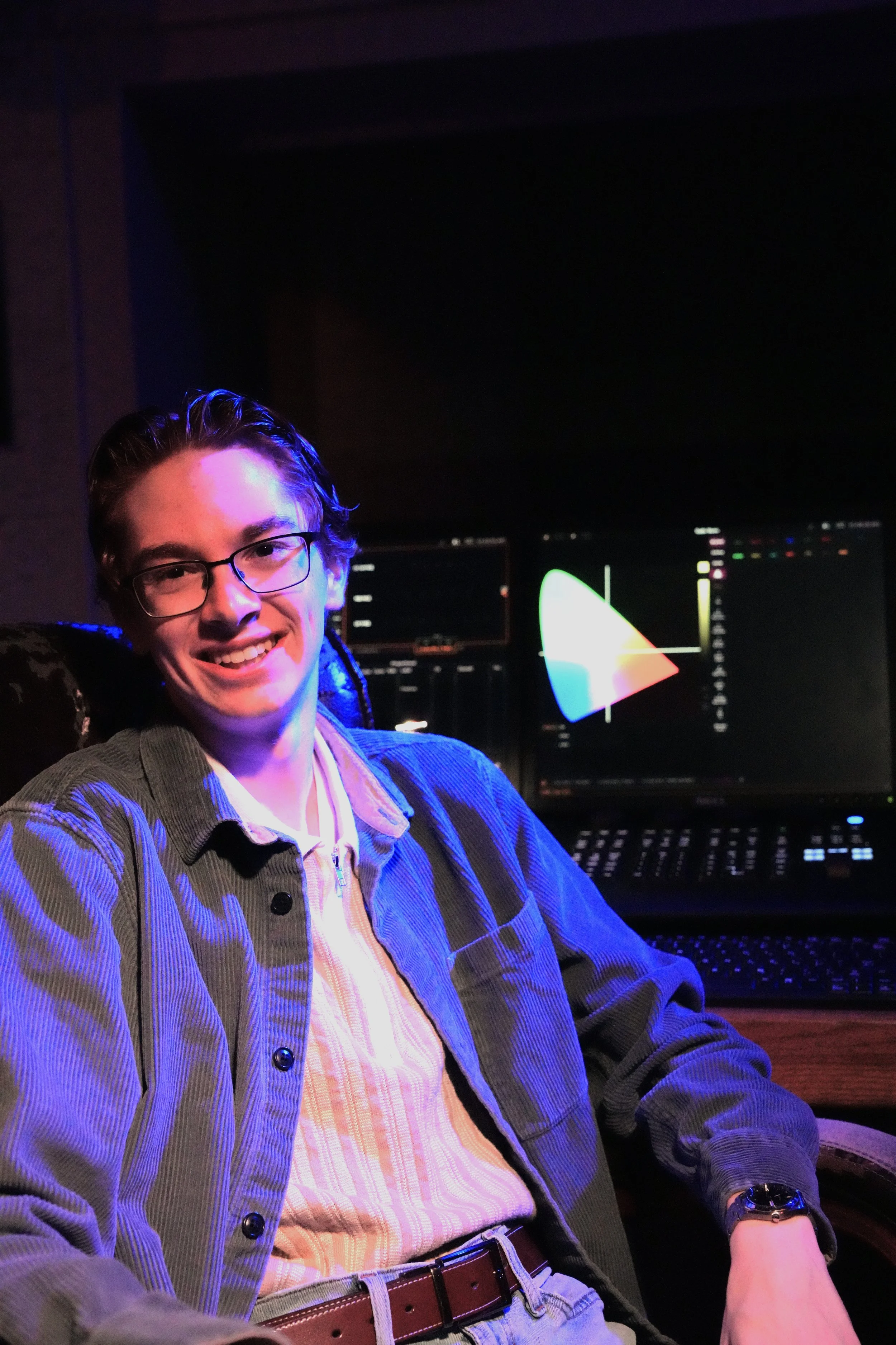 A young man with glasses smiling while sitting in front of multiple computer monitors displaying colorful graphs in a dark room.