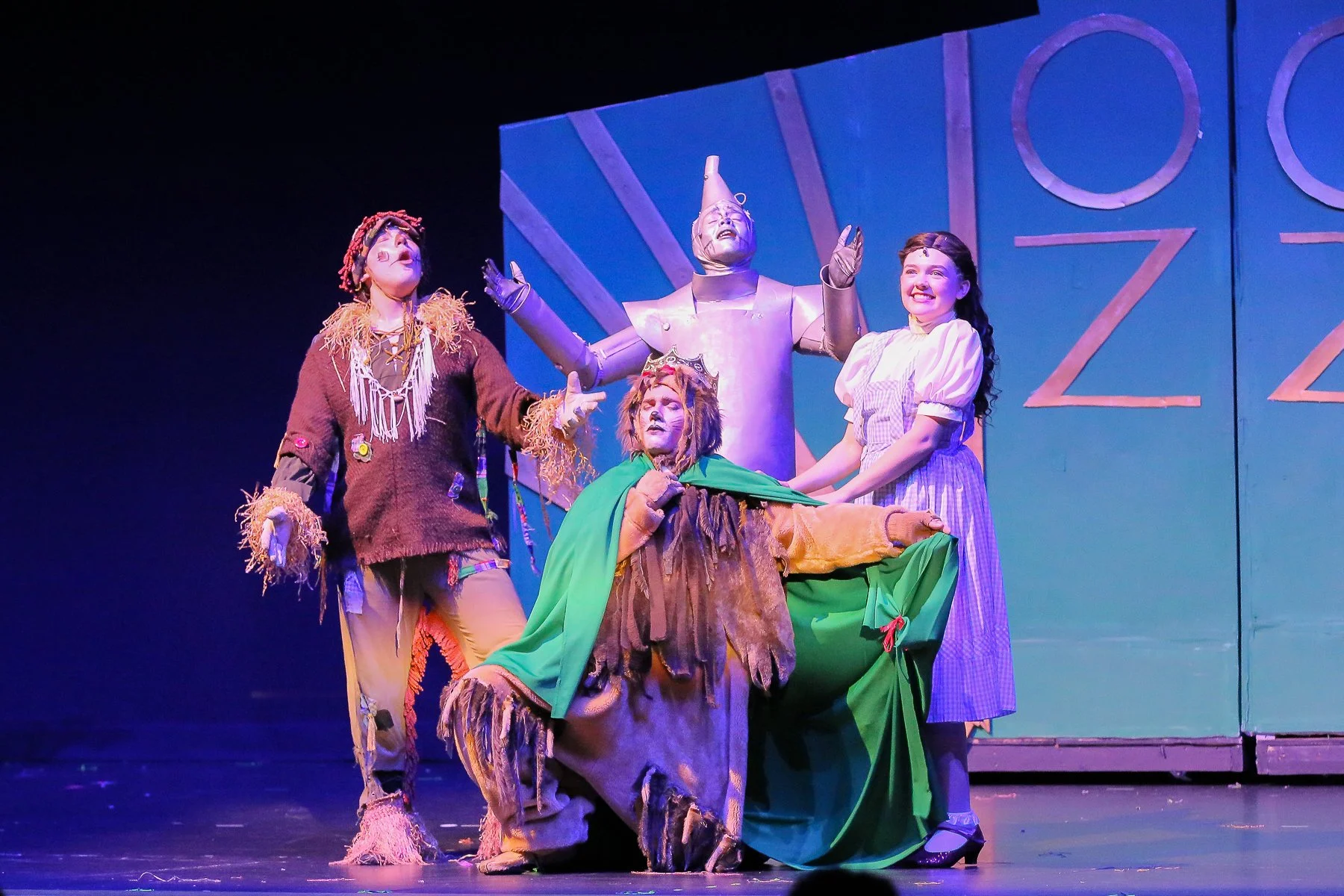 Stage scene from a musical or play with four actors in colorful costumes. One actor dressed as a scarecrow with straw accents, another as a lion, a third as a girl in a checkered dress, and a fourth as a robot with a metallic body and cone-shaped hea