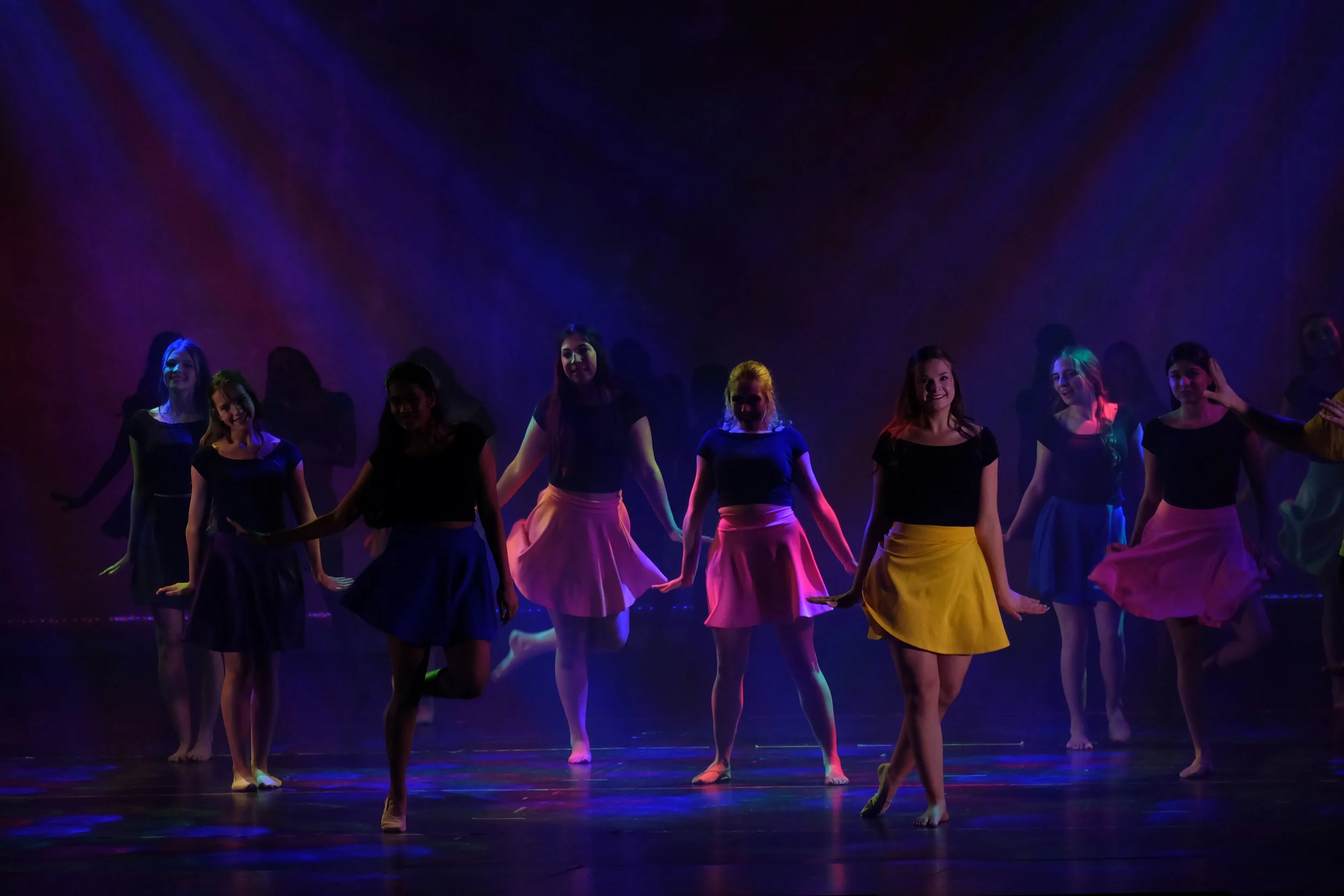 Group of young women performing a dance on stage, wearing colorful skirts and black tops, under vibrant lighting with a dark background.