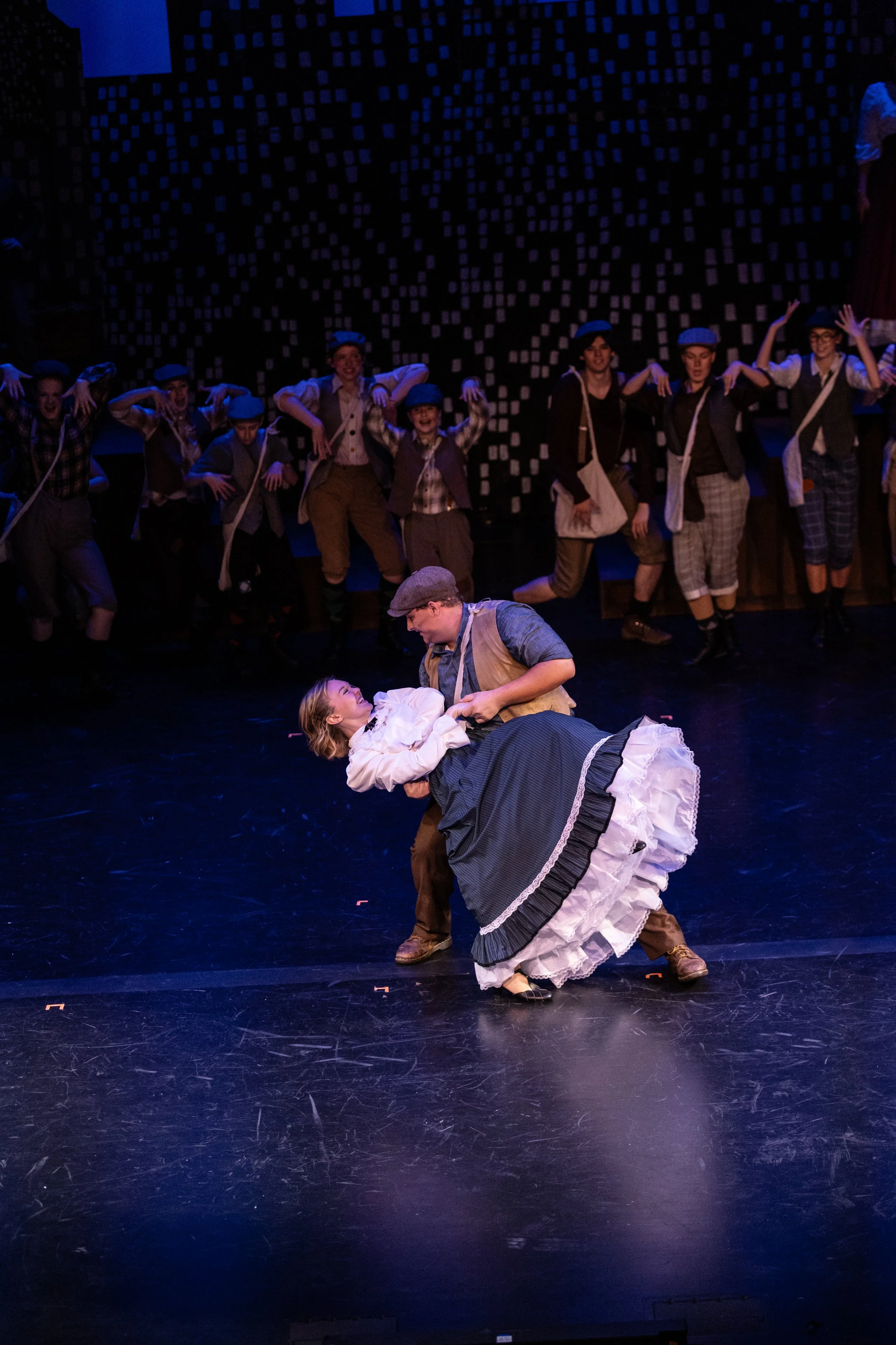 The image depicts a theatrical stage performance with actors dressed in early 20th-century costumes. Two actors are in the foreground engaged in a dance or dramatic moment, with one woman lying on the floor and the other man leaning over her. In the 