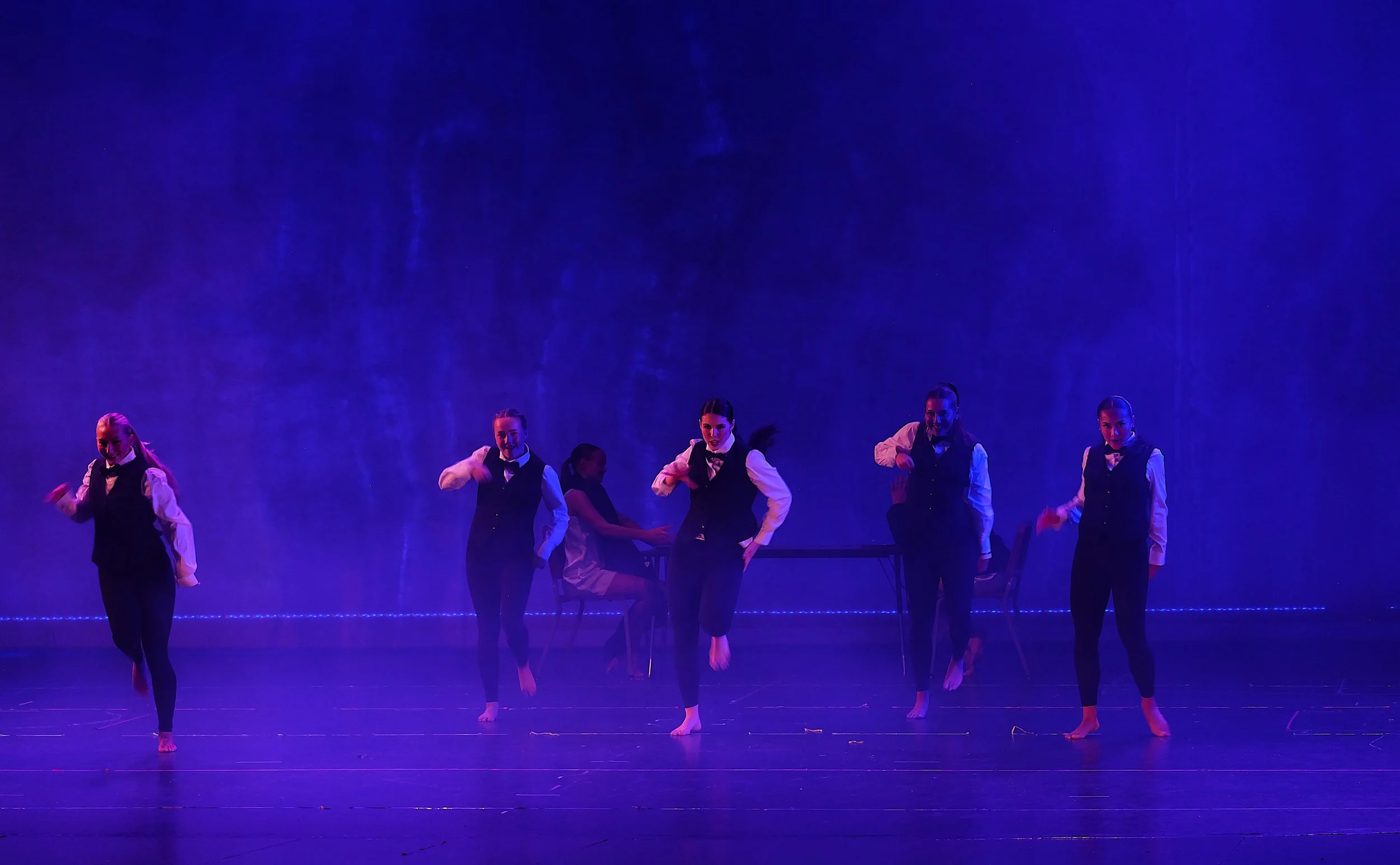 Six female dancers performing on stage under blue lighting, wearing black vests, white shirts, and black pants, with some holding arm positions and one dancer jumping.