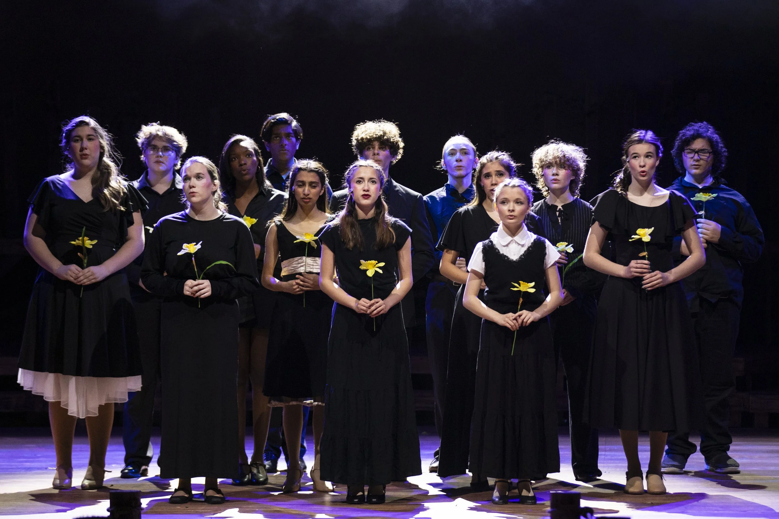 Group of young performers standing on stage, holding yellow flowers, dressed in black, during a stage production or performance.