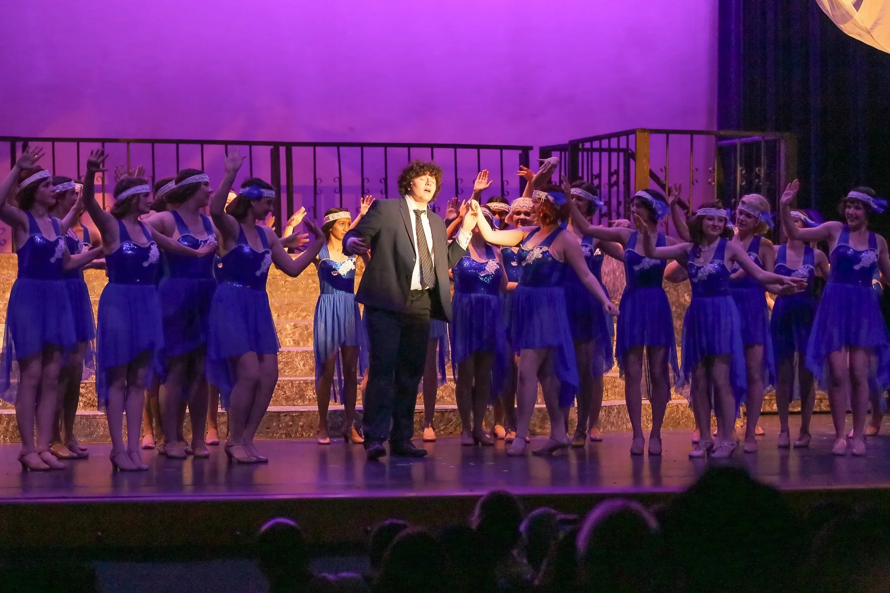 Stage performance featuring a group of young women in blue dresses with headbands and a man in a suit, all holding hands and raising them. Purple and pink lighting in the background.