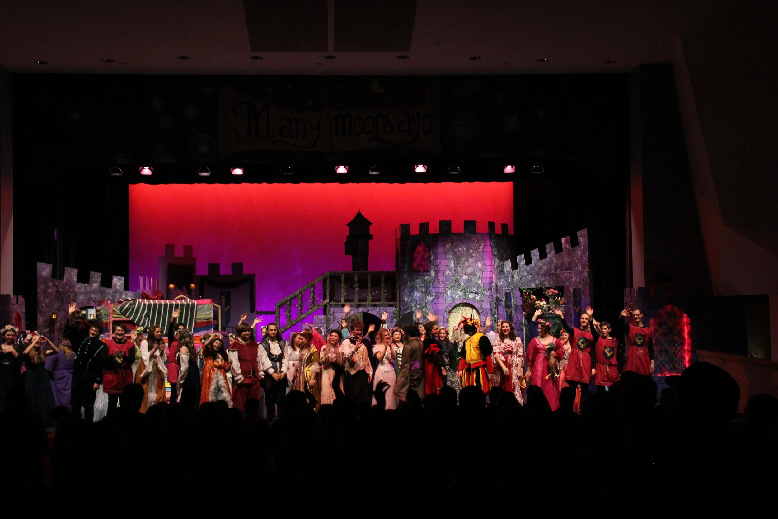 A large group of people in colorful costumes on stage, waving and smiling, with a castle-themed backdrop and stage lighting, likely after a theatrical performance or at a themed event.