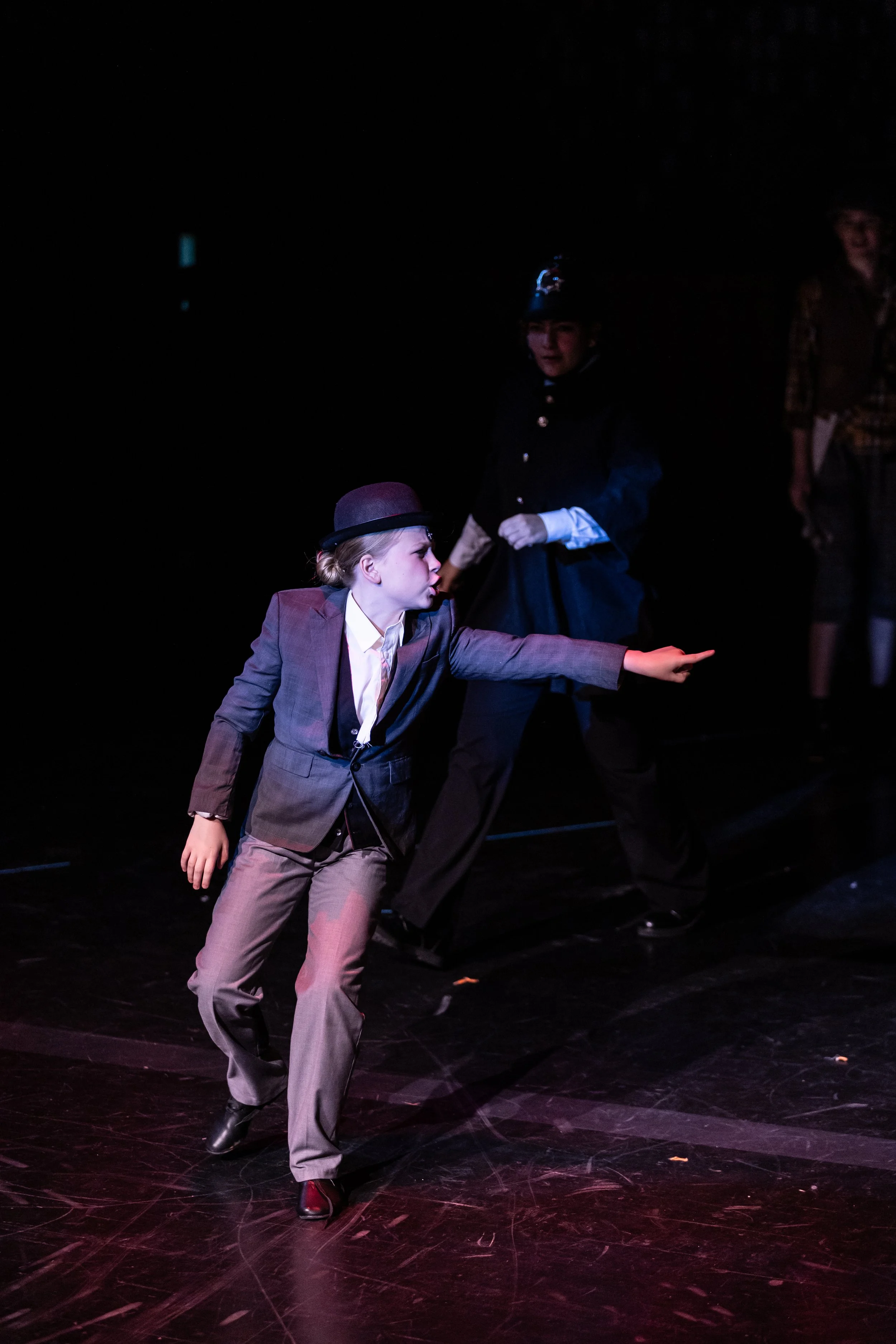 A scene from a theatrical performance with actors in costume on stage. One actor is in a gray suit and bowler hat, reaching out with one arm extended, and another actor is in a black uniform with a hat, standing behind. A third person, partially visi