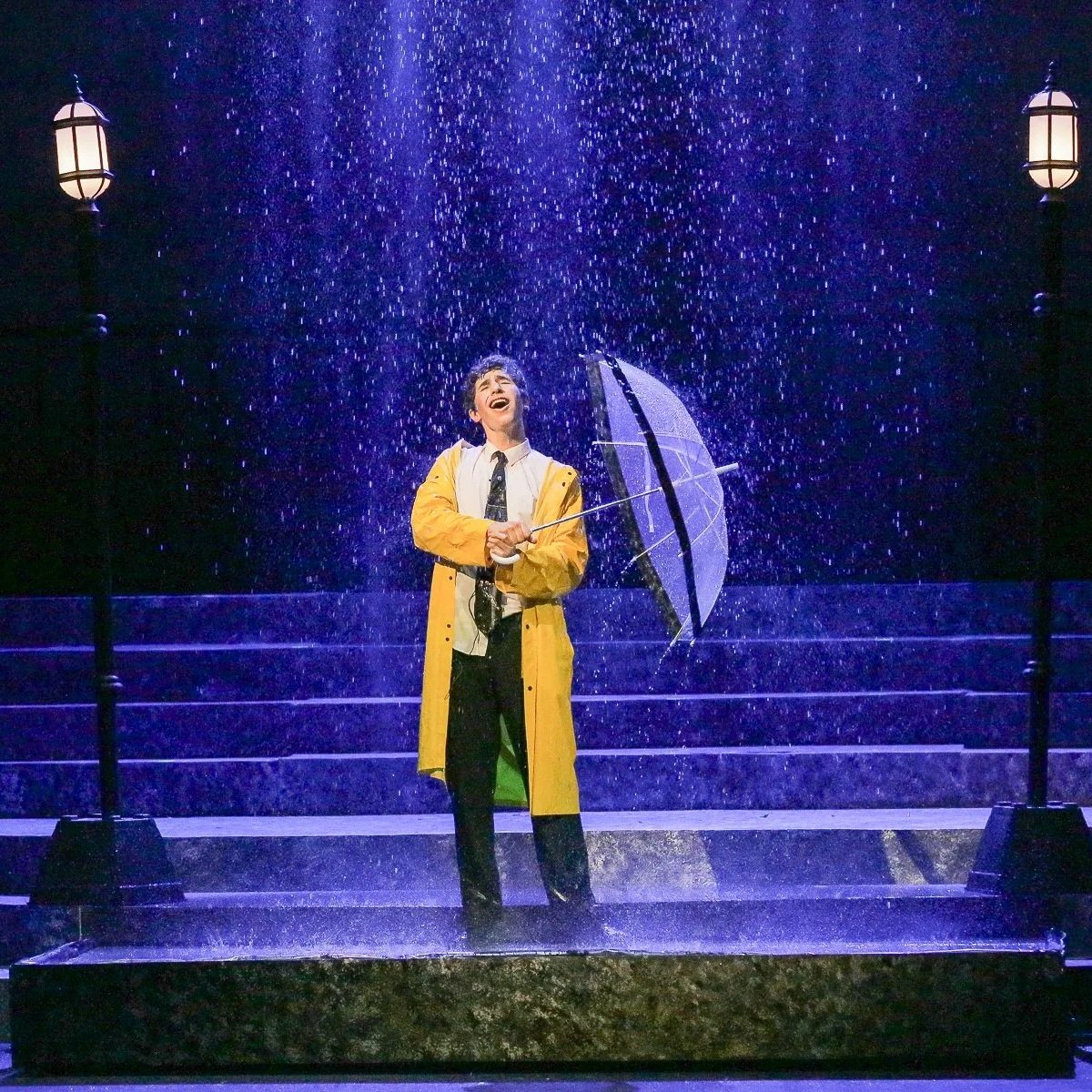 A person in a yellow raincoat holding an umbrella is standing on stairs in the rain, singing or laughing joyfully, with street lamps on either side and a dark background.
