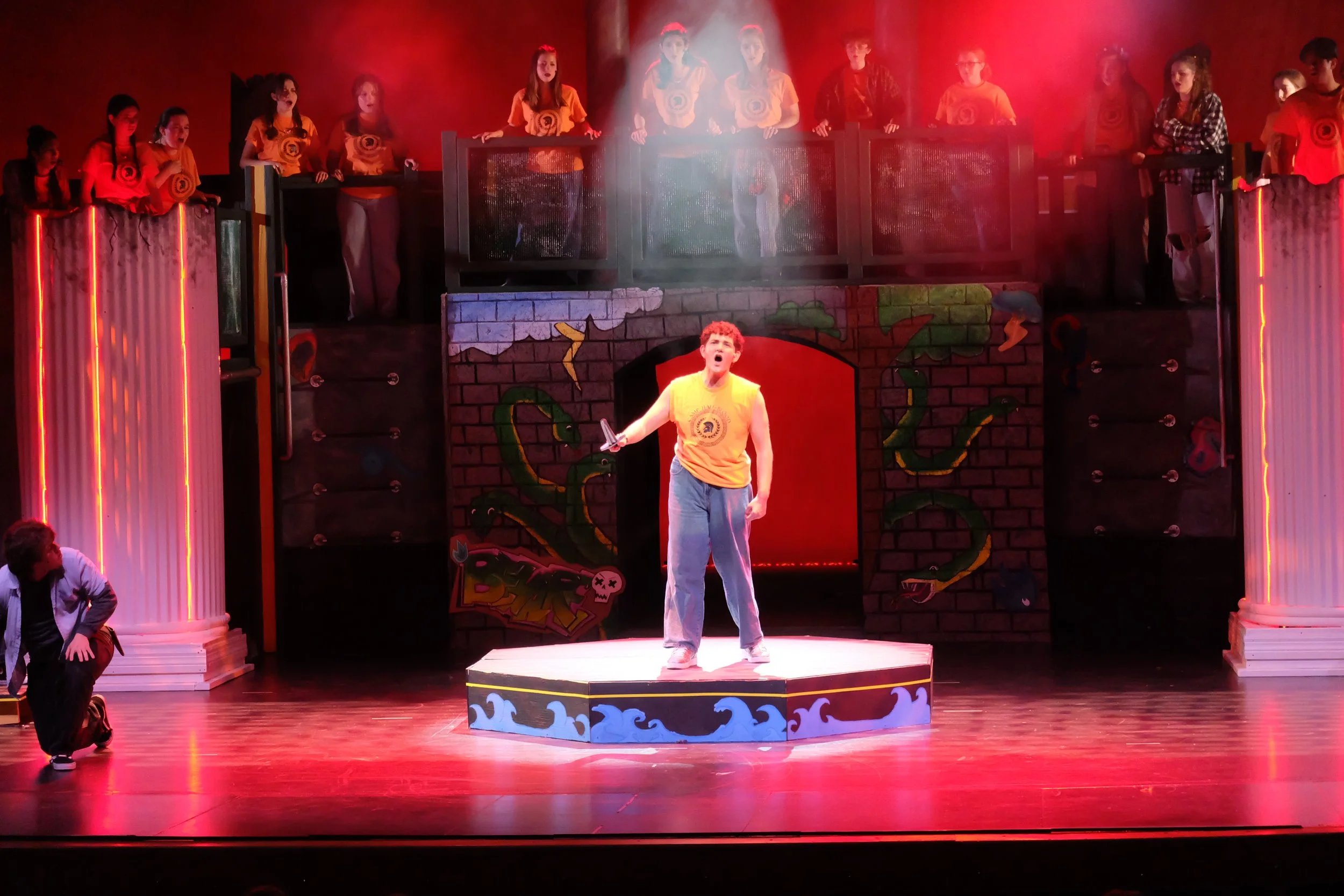 A theatrical stage scene featuring a young man in a yellow shirt and blue pants standing on a platform, holding a sword, with a group of children in matching yellow shirts and others in various costumes standing behind a balcony. The stage has colorf