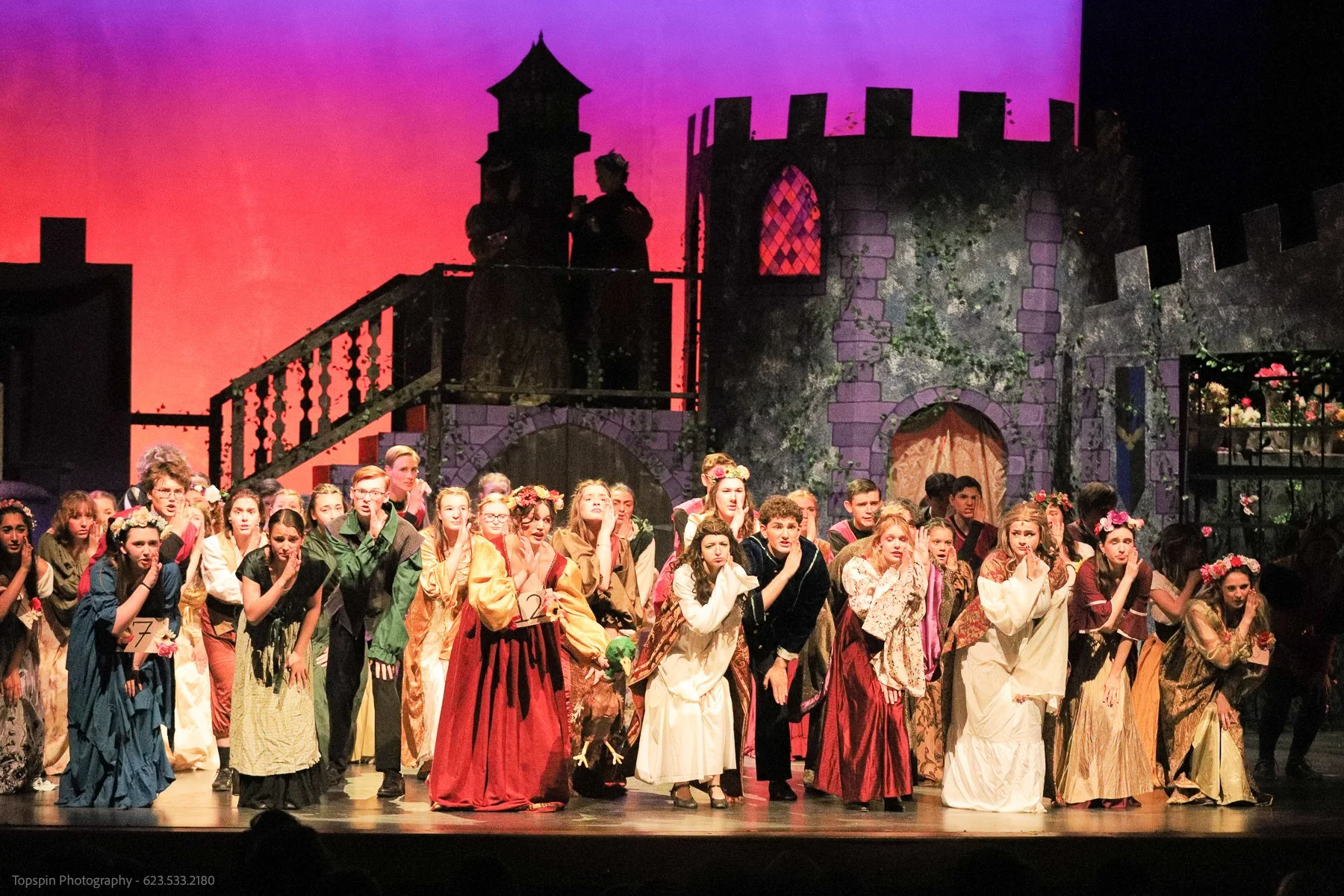 A large group of children wearing costumes and flowers on their heads perform on stage against a background of a castle with pink and purple sky. The stage appears to be for a theatrical production.