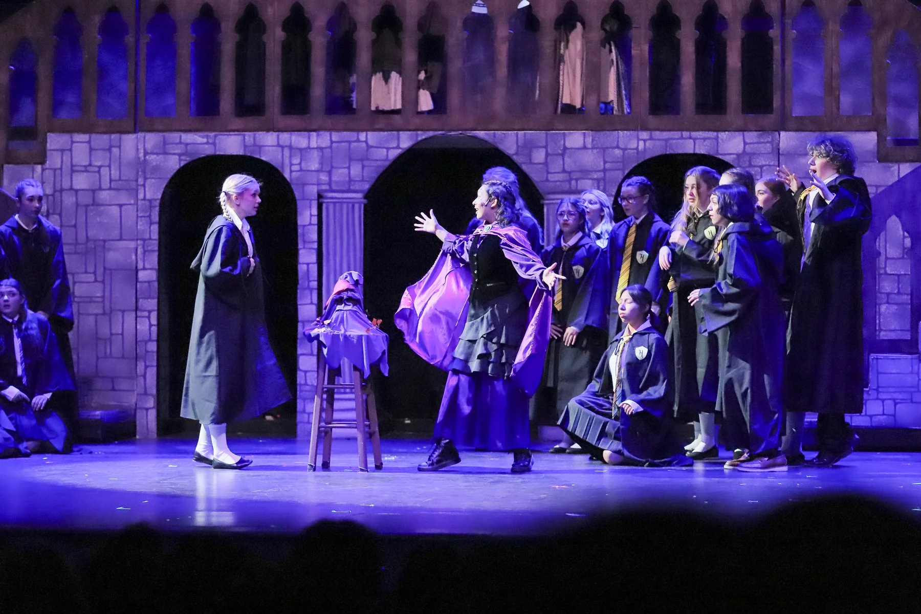 A theatrical scene with students dressed as Hogwarts students, with one woman in robes and a hat in the center, gesturing as if teaching or telling a story. The stage has a castle-like backdrop with arched windows and a balcony, and the students are 