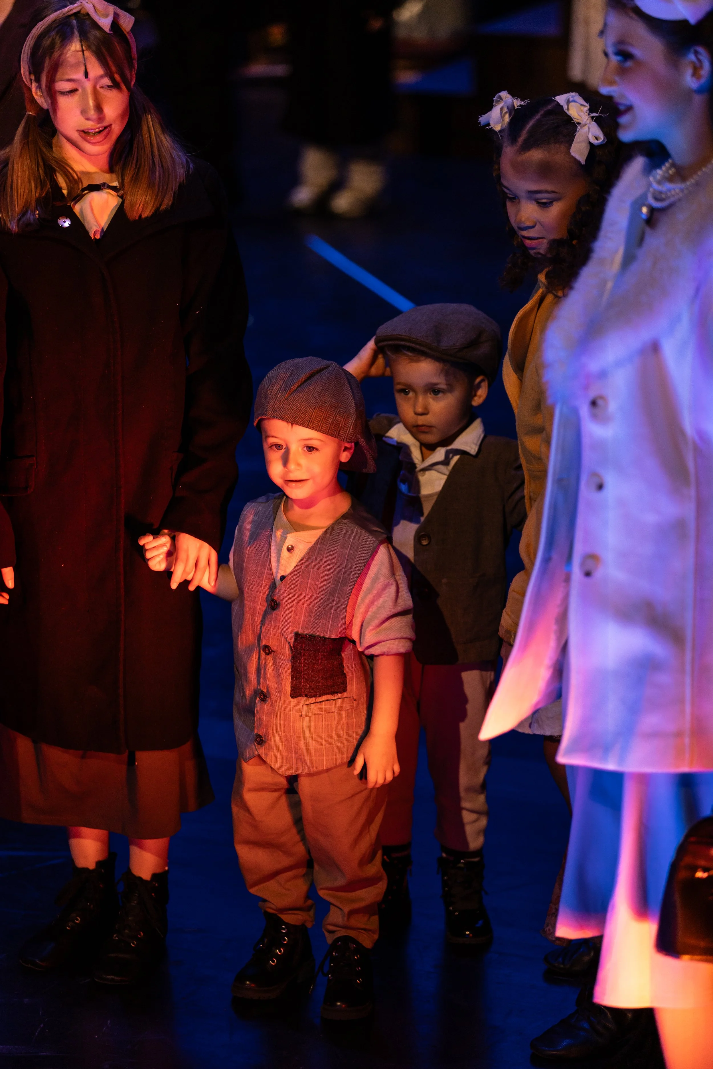 A group of children dressed in vintage costumes, holding hands and standing on a dark stage with colorful lighting.