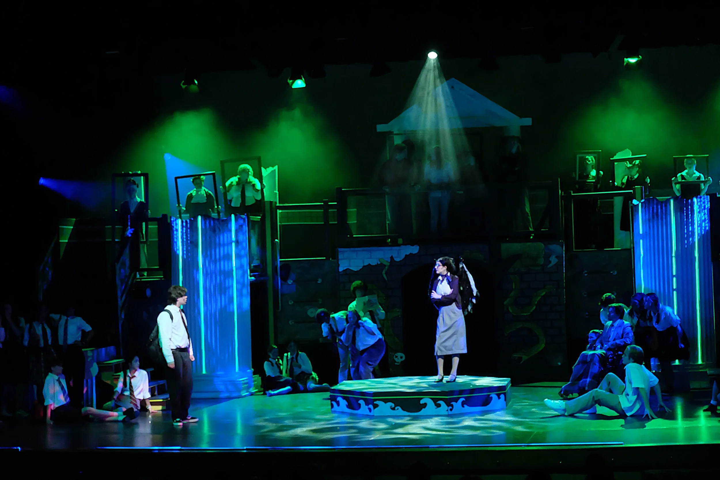 A theatrical stage scene with multiple performers, some standing and some sitting, under green and blue lighting. The stage features a pirate ship structure in the background and various stage props, with some performers dressed in school uniforms an