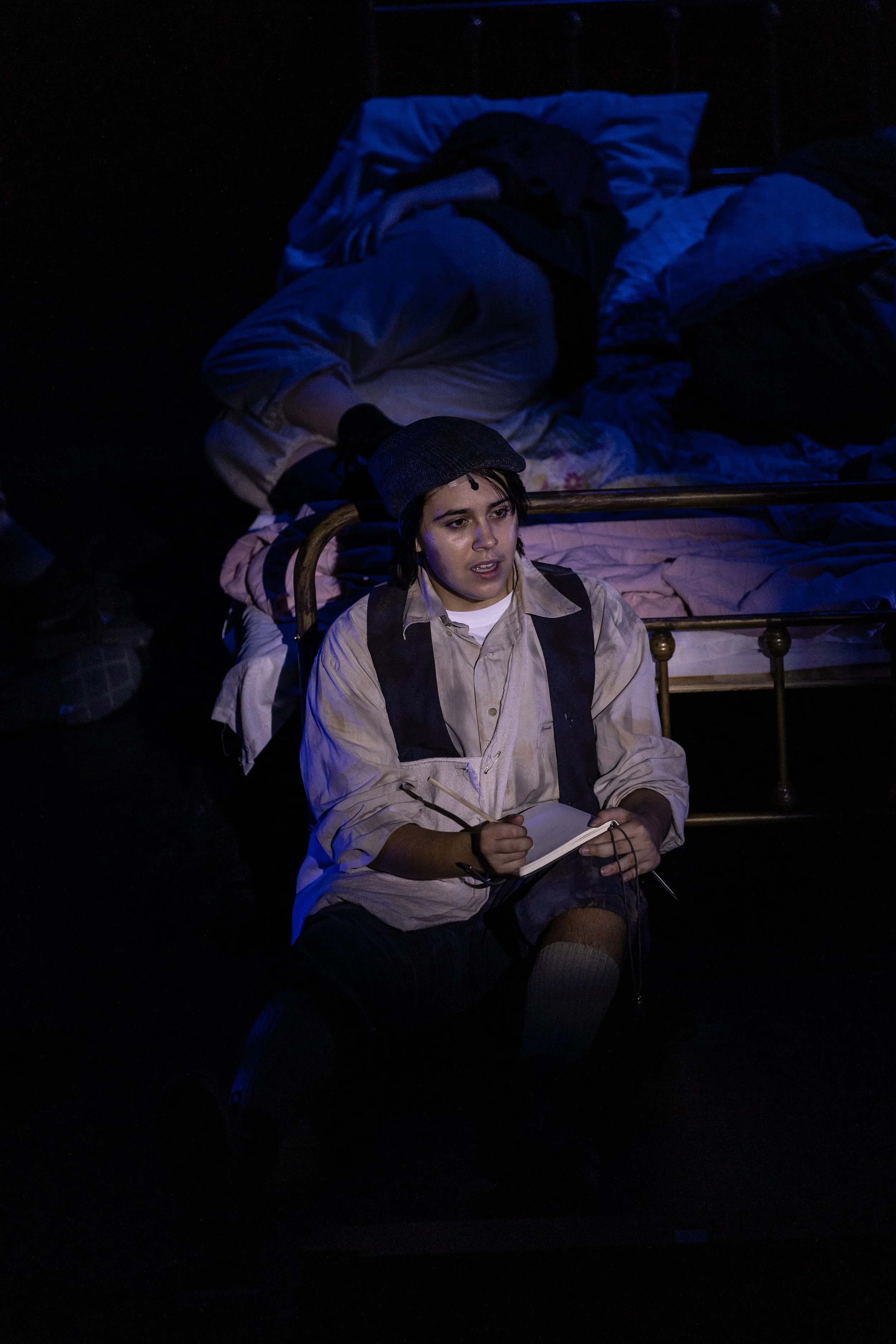 A young man sitting on a chair with a notebook, wearing a cap, with a person lying on a bed in a dark room, illuminated by dim blue lighting.