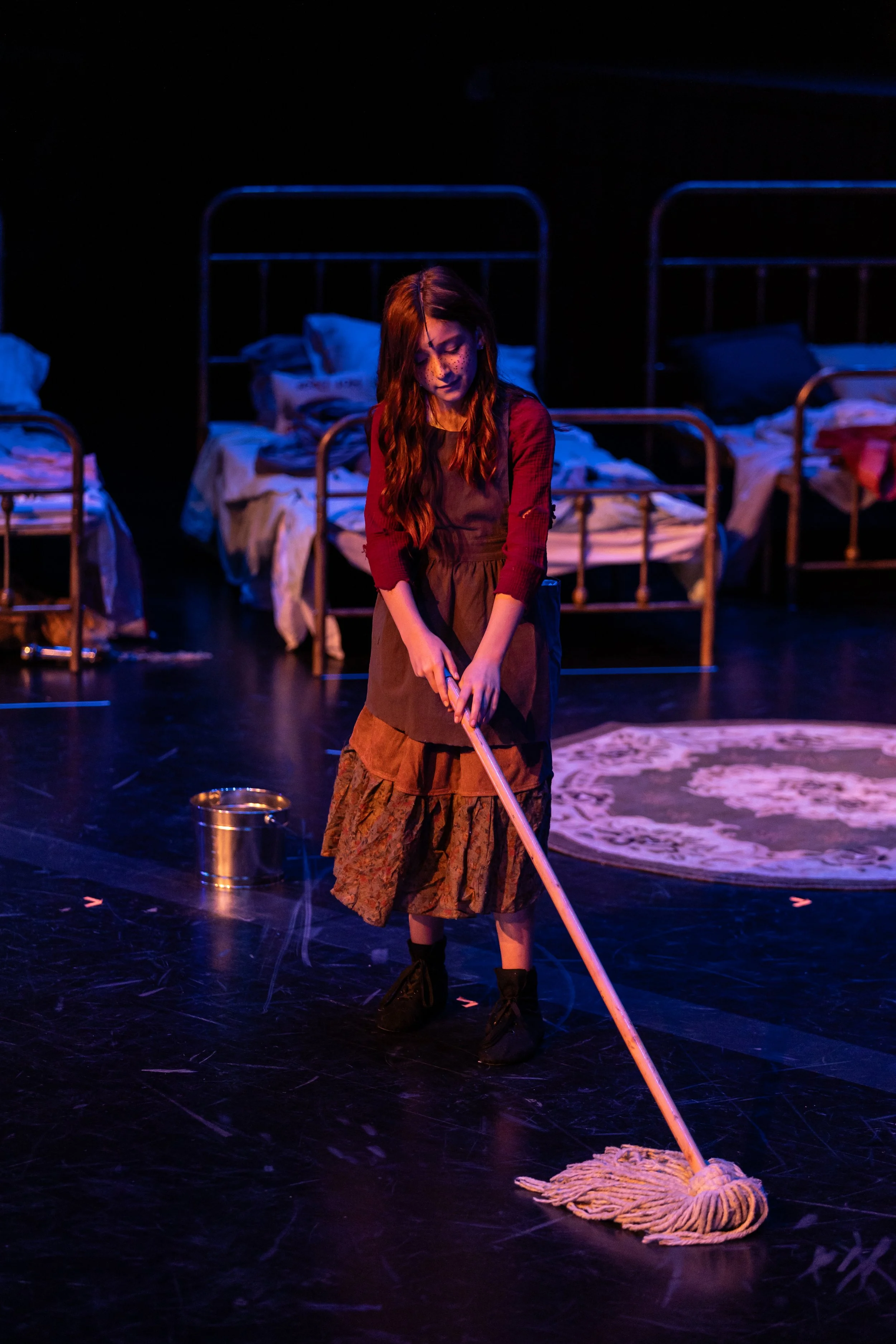 A woman with long brown hair, wearing a brown skirt and a red long-sleeve shirt, stands on a stage mopping the floor with a mop and a bucket nearby. In the background, there are three metal beds with disheveled sheets and blankets, in a dimly lit sce
