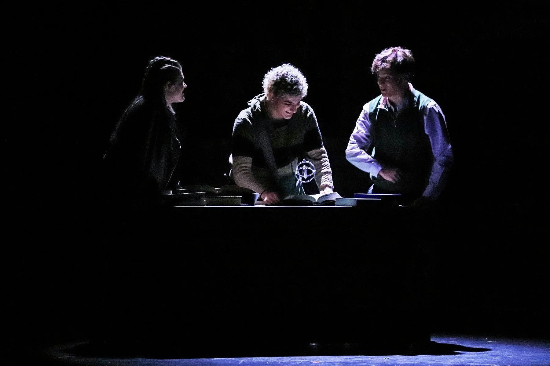 Three young people gathered around a table with an open book, illuminated by a single light in a dark setting.