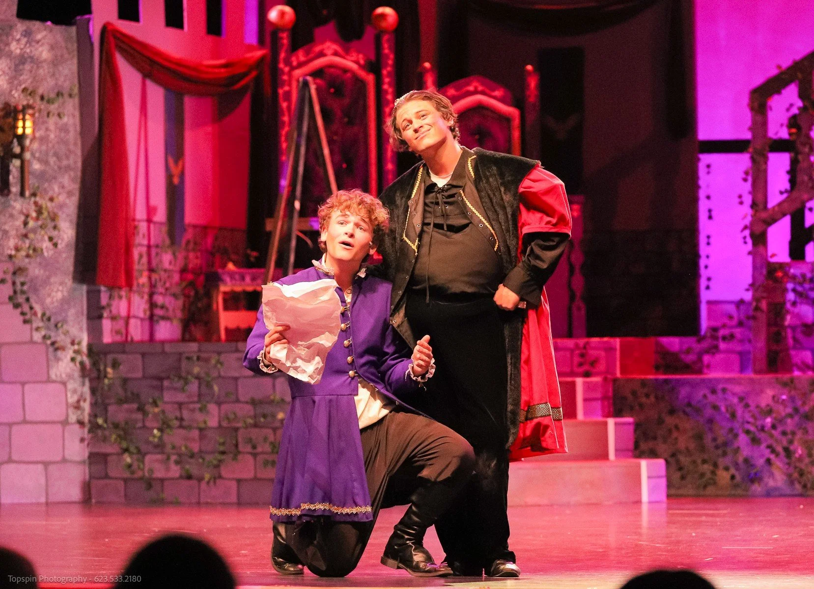 Two actors on stage during a theatrical play; one kneeling, holding a paper bag and an expression of surprise, the other standing with a confident smile, dressed in costume with a black coat and red sleeves, set against a medieval castle backdrop.