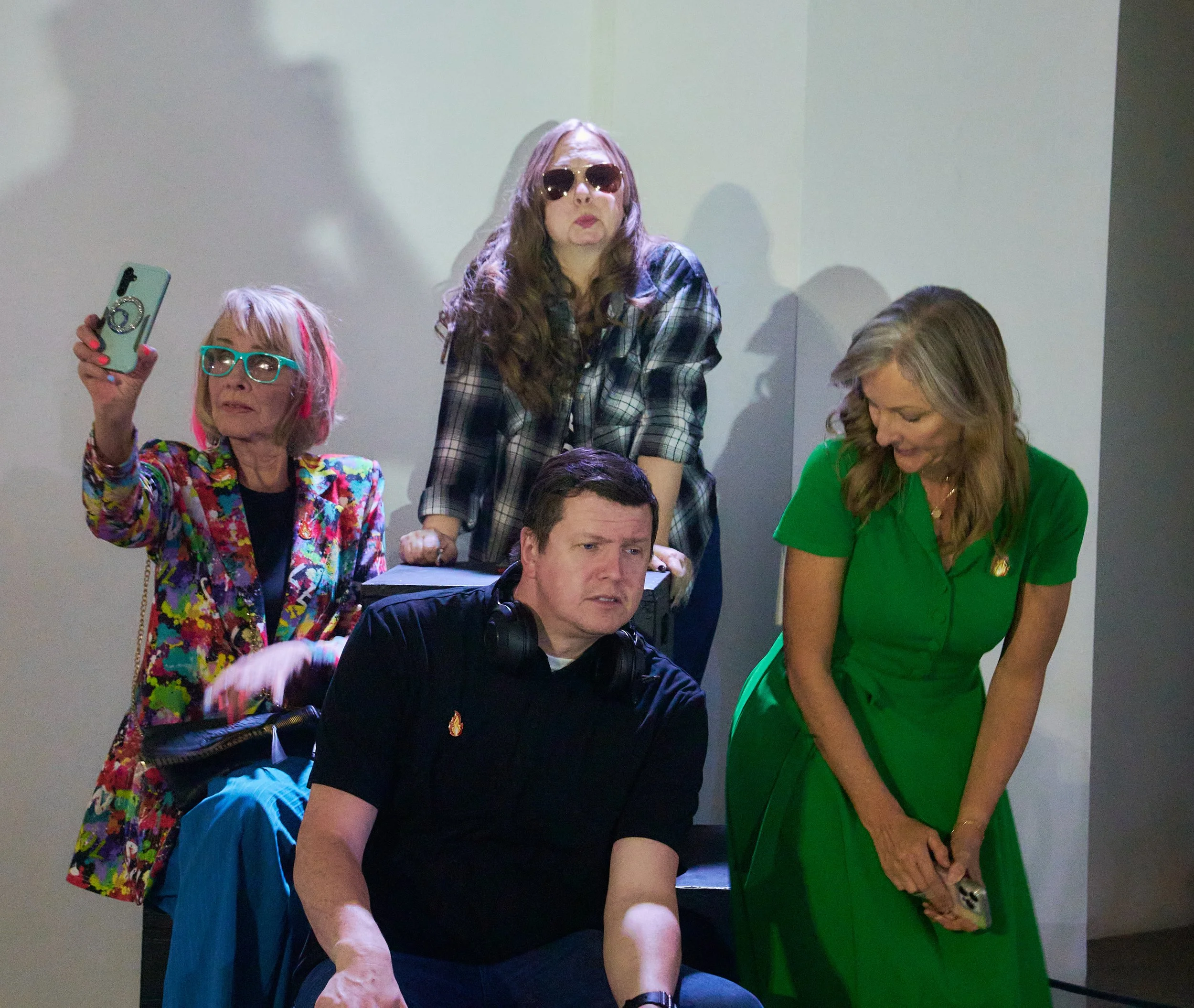 A group of people, mostly women, gather around a man seated in a chair, with one woman standing behind him. The woman on the left wears colorful clothing and neon glasses, holding a phone, and the woman on the right is dressed in green. The woman sta