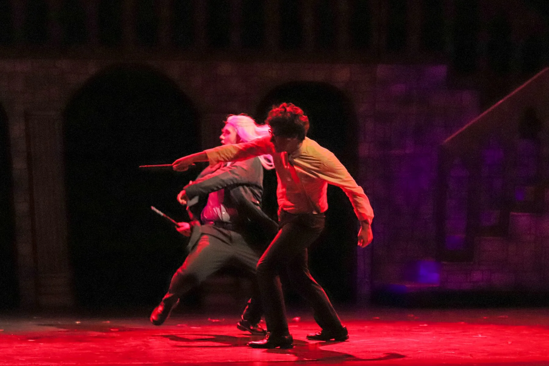 Three performers in dark clothing engaged in a physical dance or theatrical act on a dark stage illuminated with red and purple lighting.