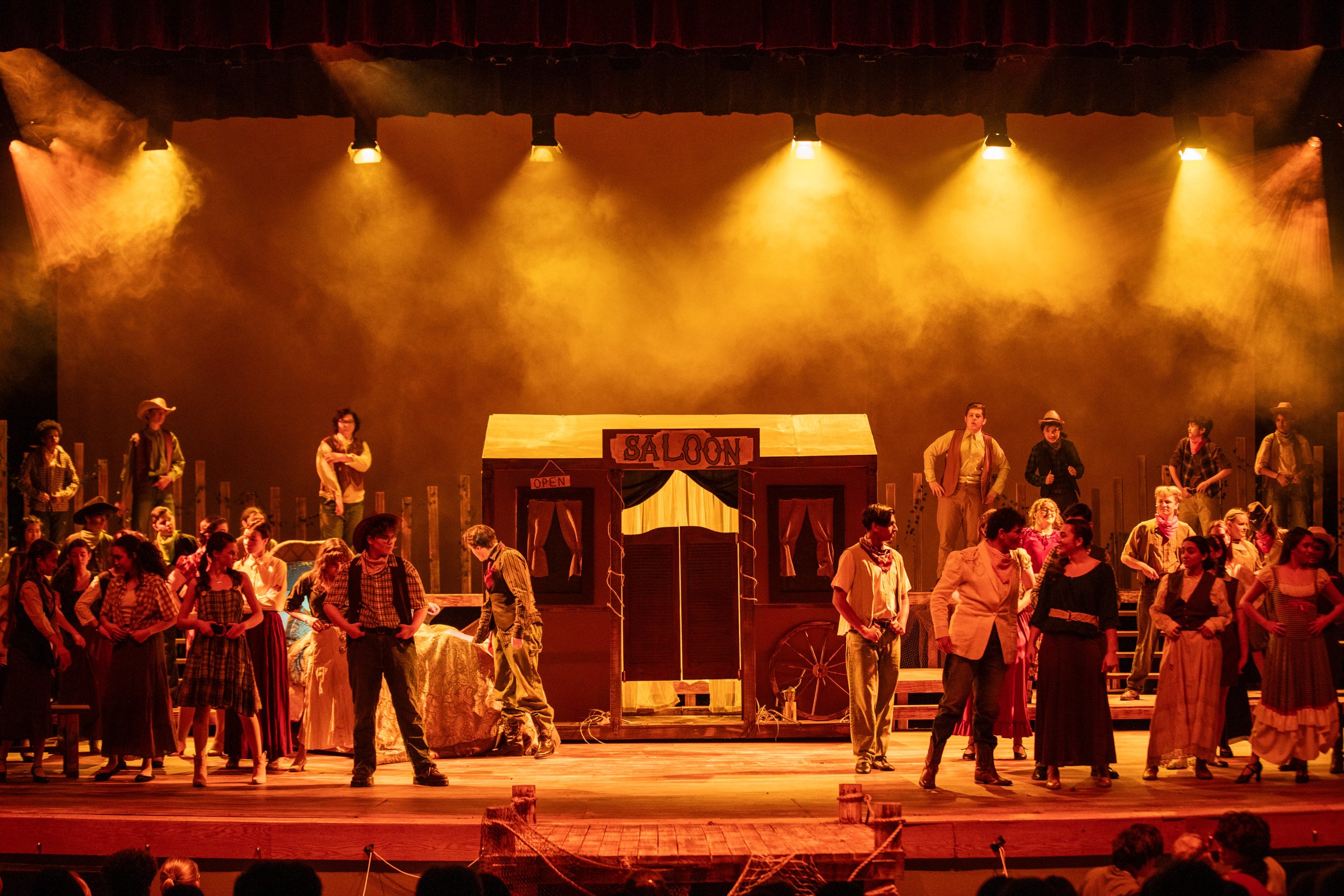 A large stage with yellow and orange lighting, featuring a theatrical set resembling a saloon with a wooden facade and a sign that says 'SALoon.' There are many actors dressed in cowboy and western-style costumes, some standing and others sitting, wi
