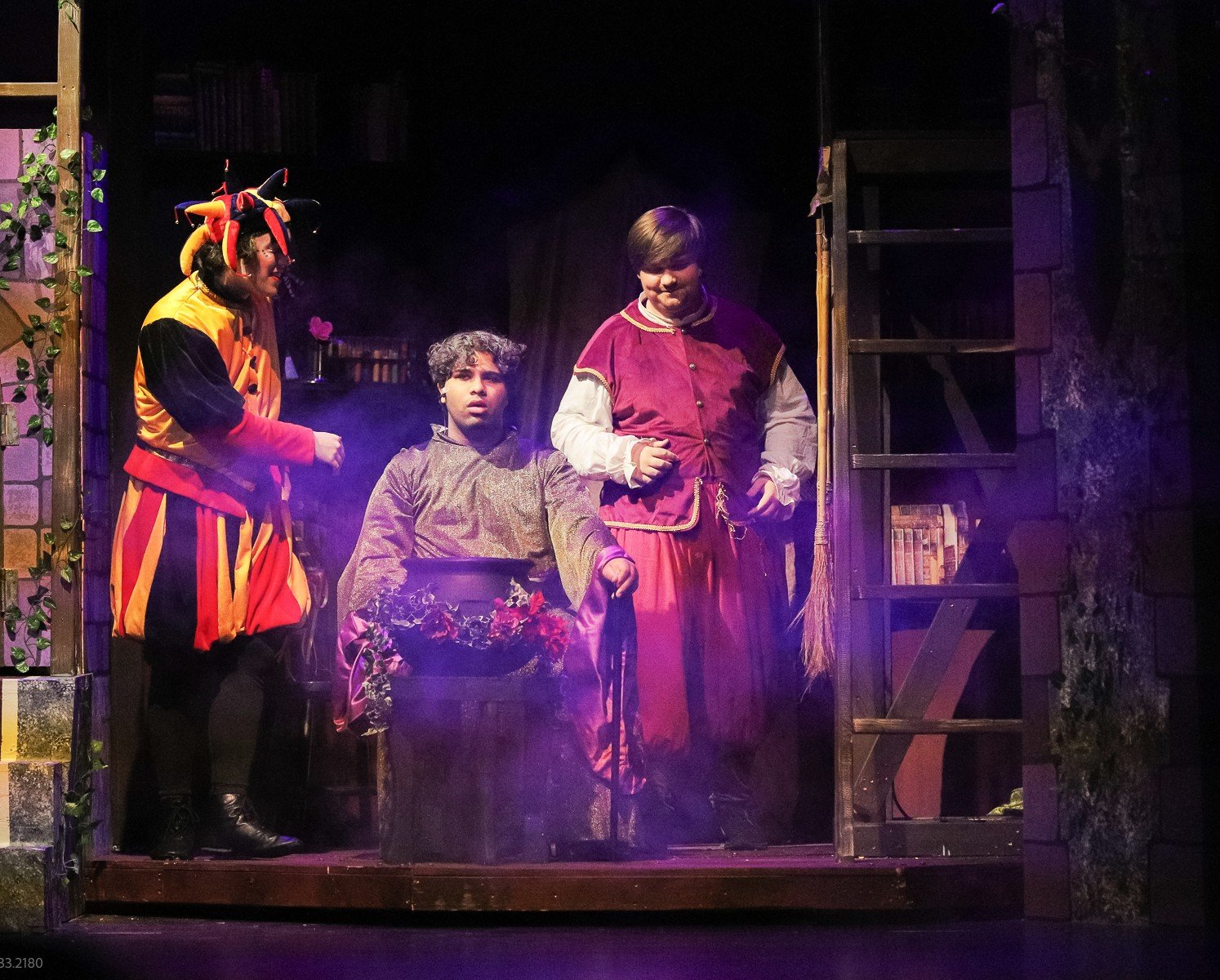 Three actors on stage in a theater play, with one dressed as a jester or clown, and two others in medieval or fantasy costumes, standing near a pot with flowers, set against a dark backdrop with stairs and bookshelves.