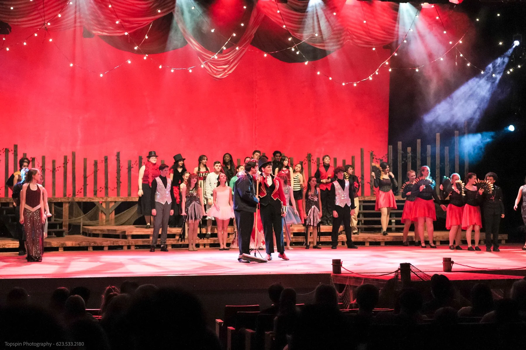 A large theatrical stage with a red backdrop and string lights, showing many performers in colorful costumes, some holding fans, and a few in circus or showgirl outfits.