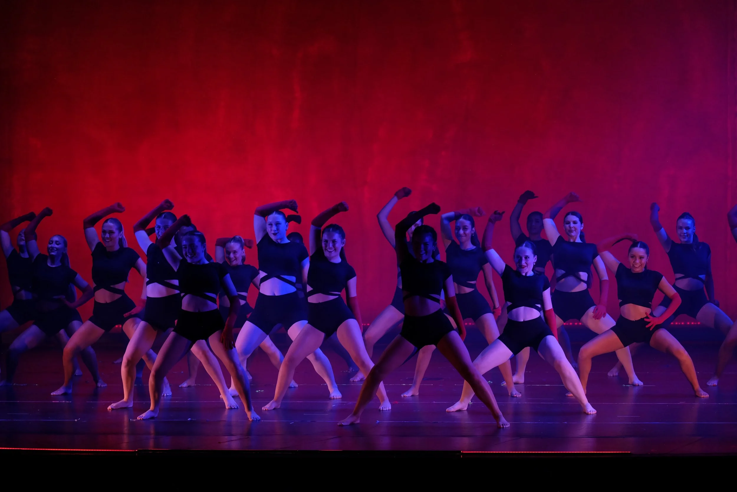 A group of female dancers in black costumes performing on stage with a red and purple background.